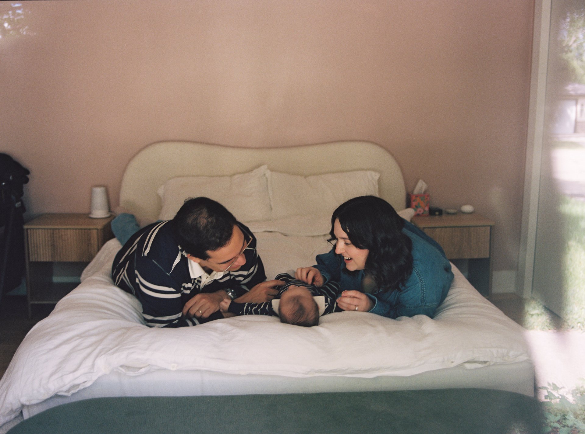 A family of three, including a man, woman, and baby, playing together on a bed in a cozy bedroom.