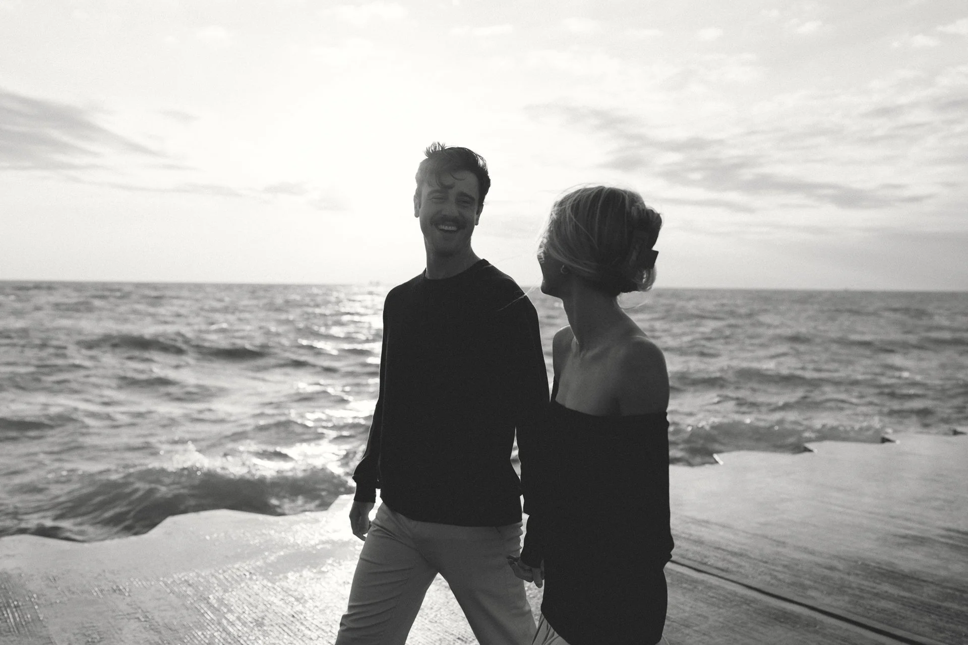 A smiling man and woman holding hands on a beach at sunset, with the ocean in the background.