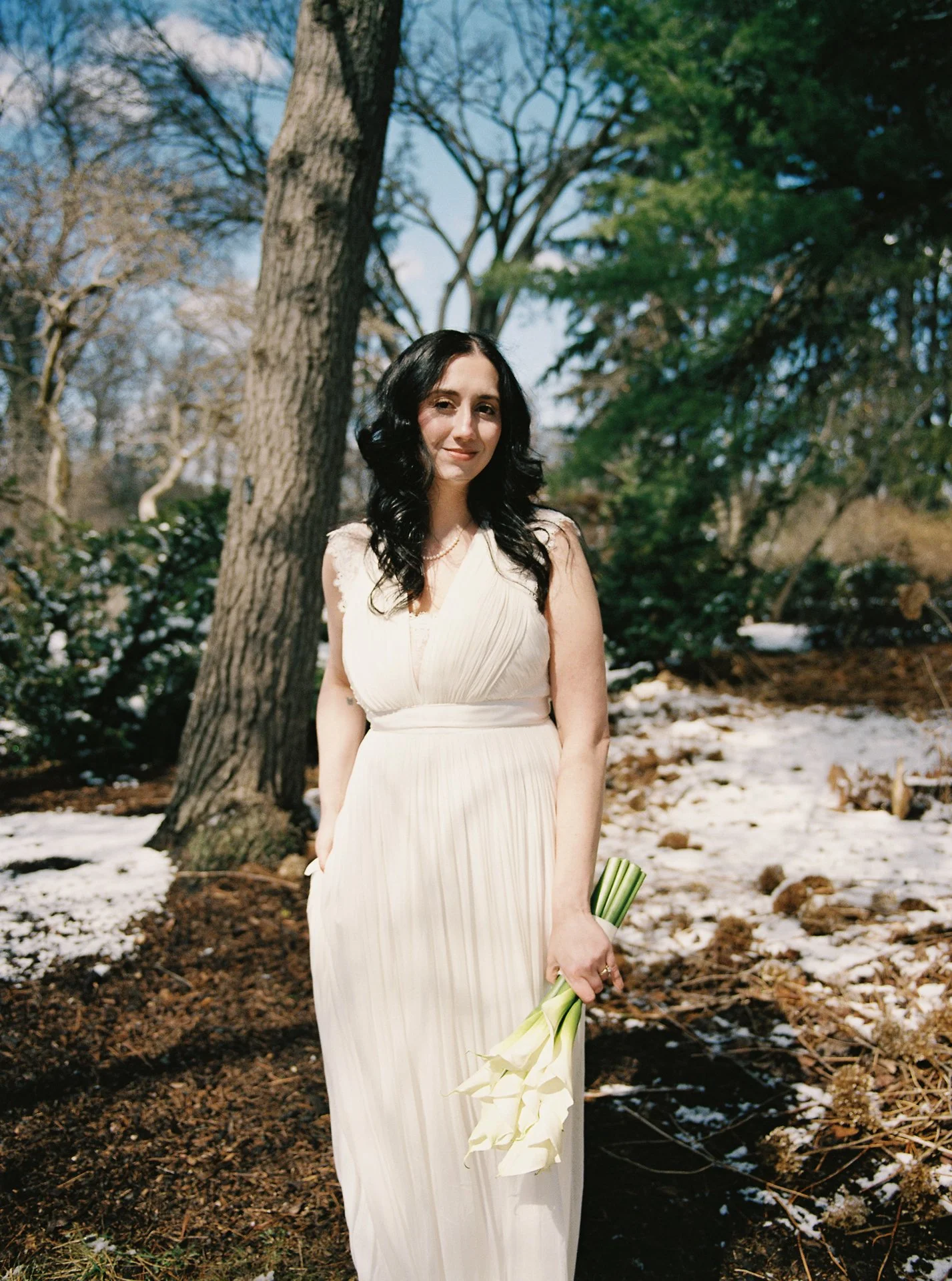 A woman in a white dress holding a bouquet of calla lilies, standing outdoors with trees and patches of snow on the ground.