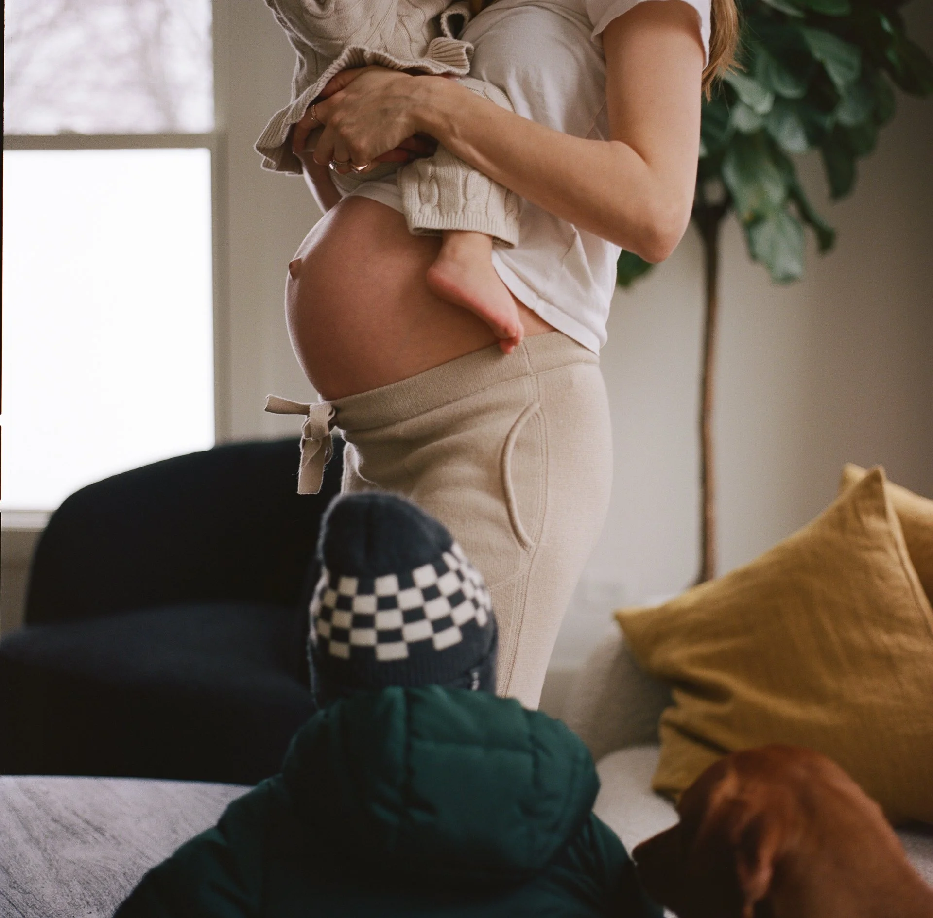 A pregnant woman holding a young child while seated in a living room, with a dog nearby and a window in the background.