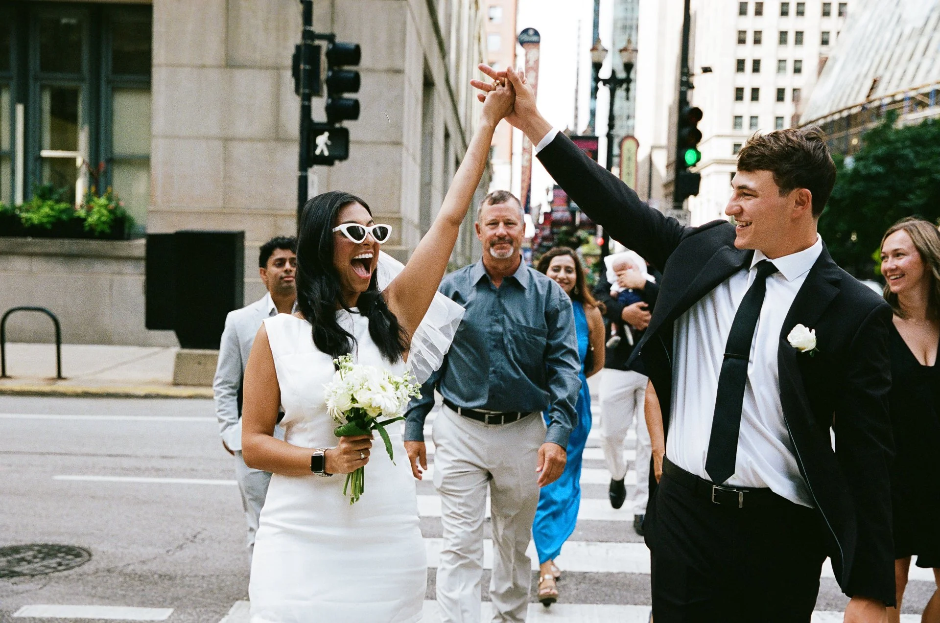 A wedding couple celebrates by holding hands and smiling on a city street as friends and family watch.