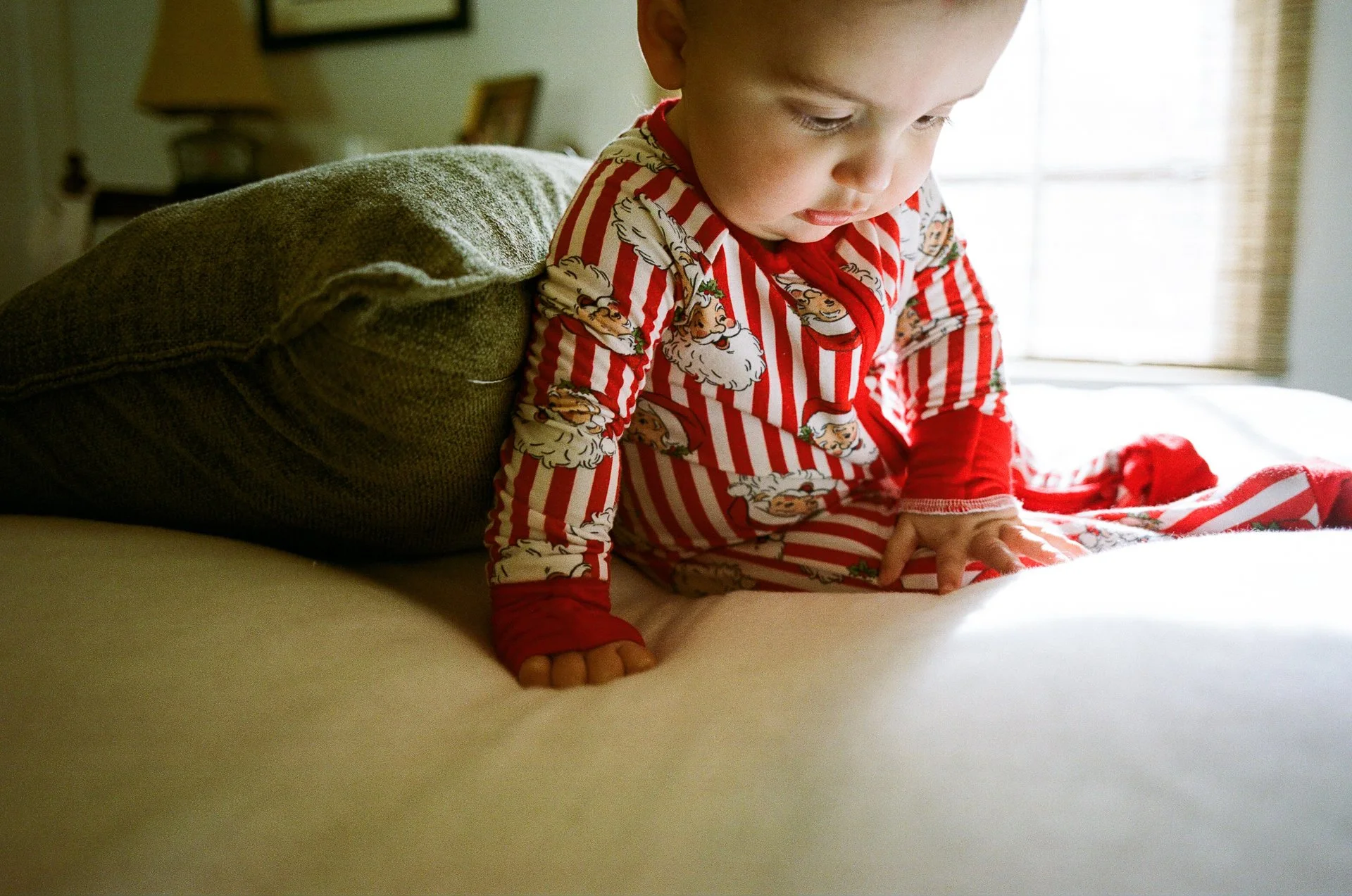 Young child wearing red and white striped pajamas with Santa Claus print, kneeling on a bed, leaning forward with hands on the mattress, in a bedroom with a window and lamp in the background.