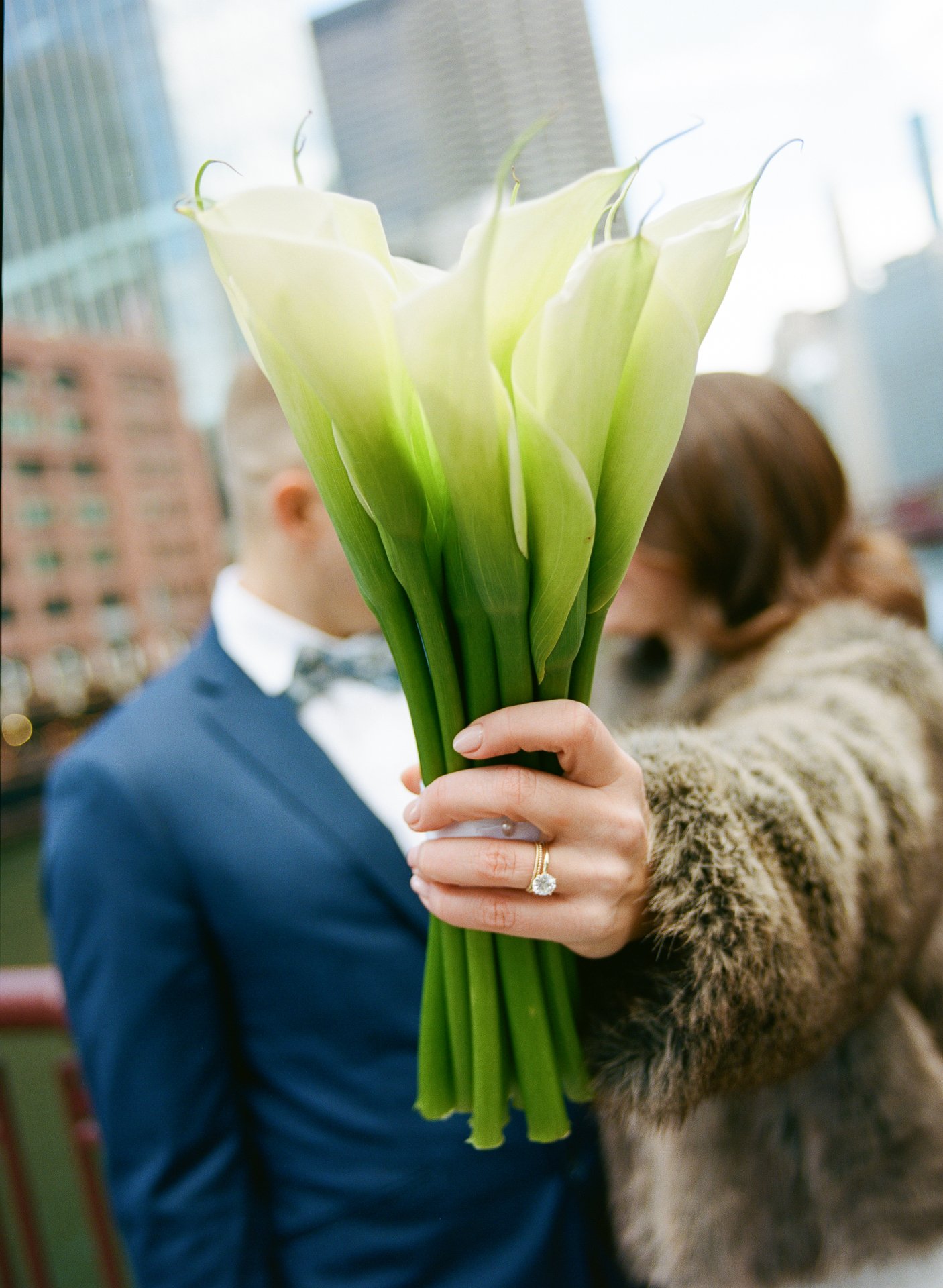 A woman holding a bouquet of white calla lilies in front of her face, with a man in a suit and bow tie blurred in the background on an urban rooftop.