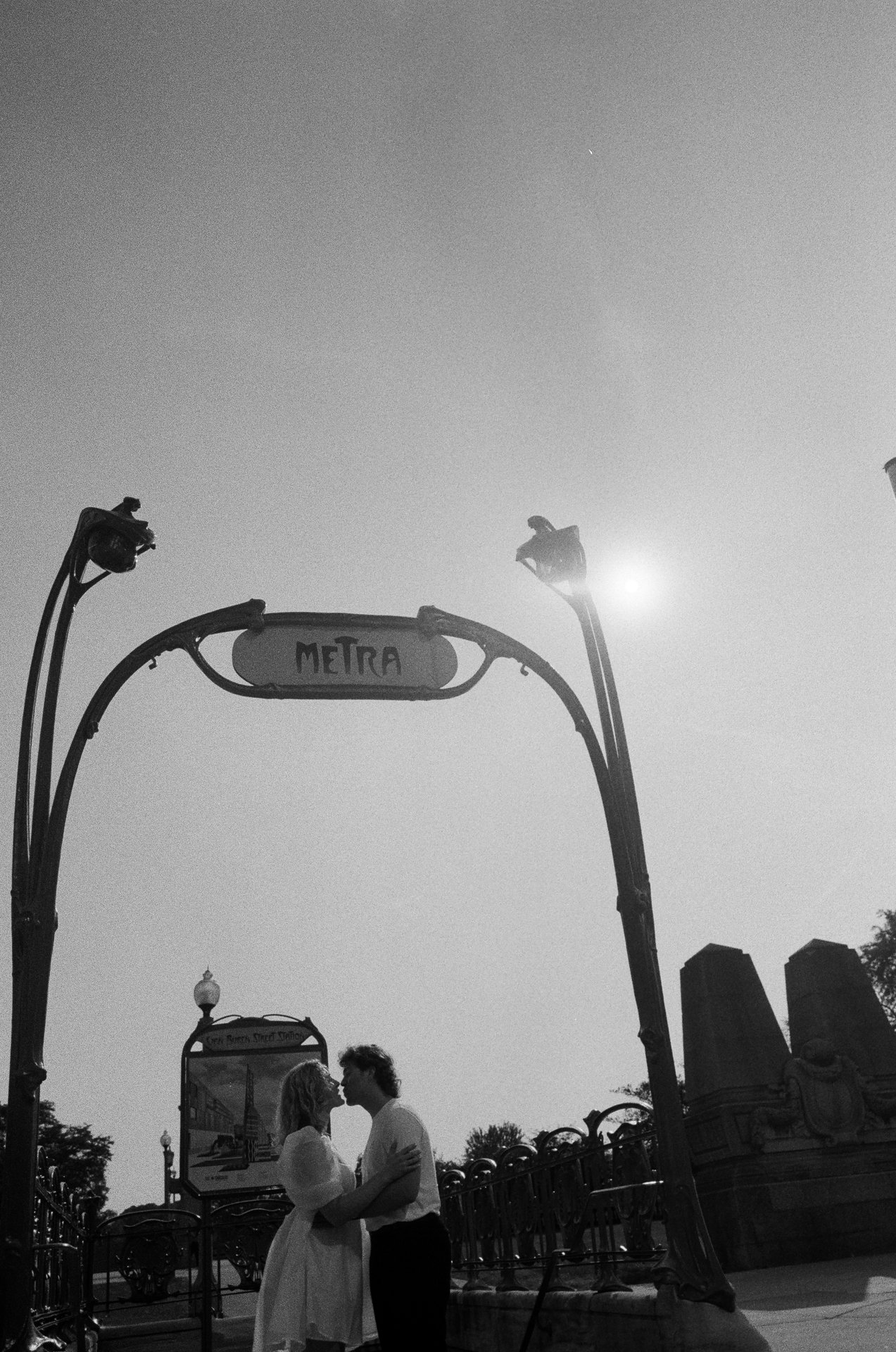 A couple kissing under an ornate Metro sign, with a clear sky and Sun in the background.