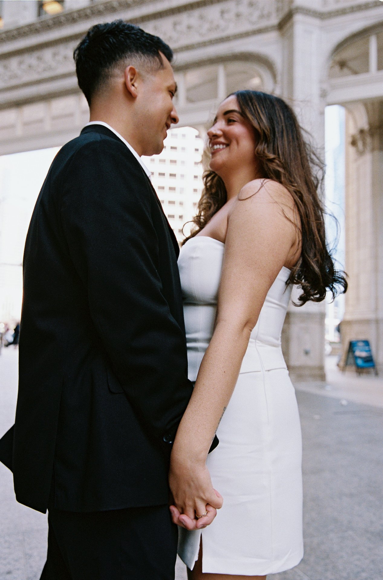 A couple holding hands and smiling at each other outdoors near a stone archway, with city buildings in the background.