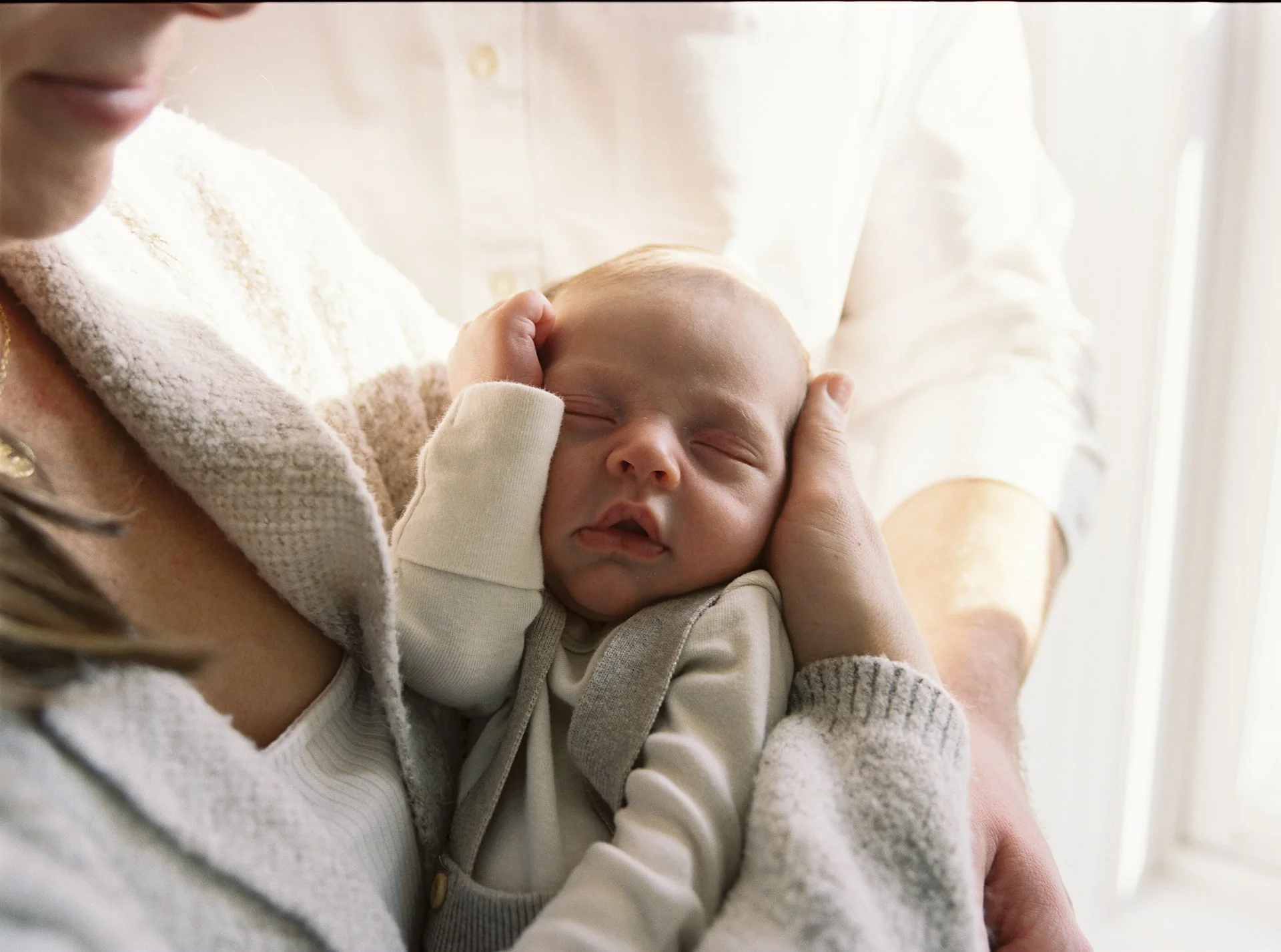 A baby is being held by an adult, appearing to sleep peacefully with eyes closed and mouth slightly open, with the adult gently supporting the baby's head and shoulder near a bright window.