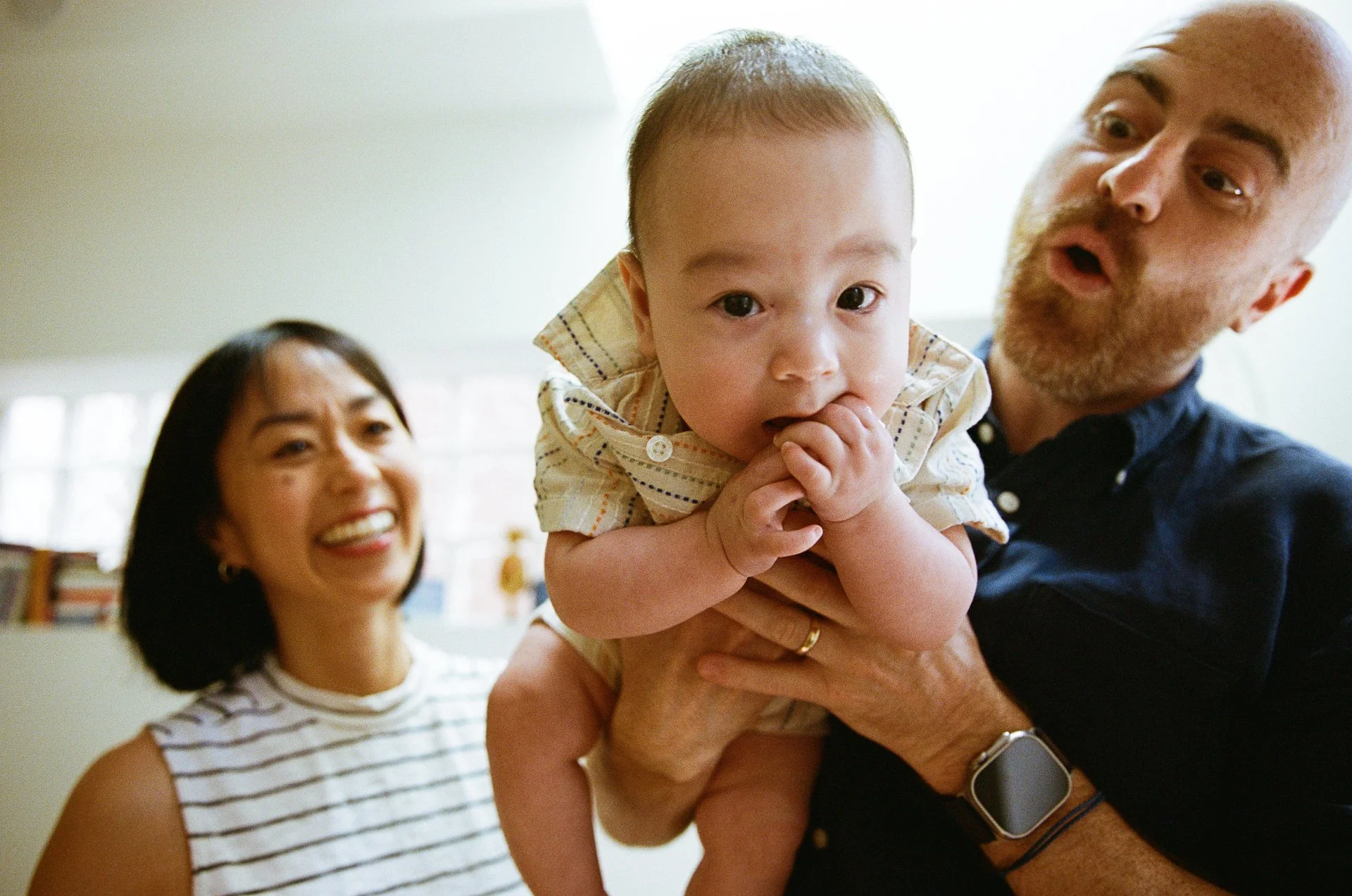 A family with a woman, a man, and a baby in a bright room. The woman is smiling, the man is holding the baby, who has a curious expression and is licking fingers.