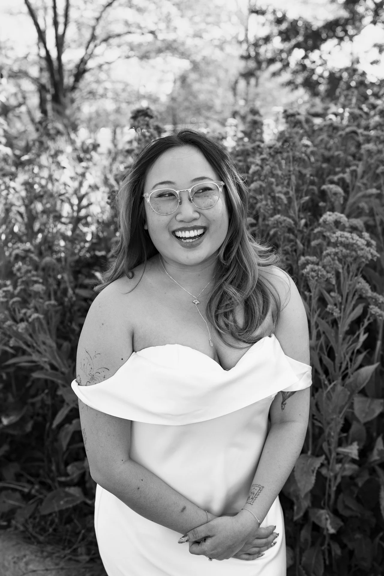 A woman in a white off-the-shoulder dress, smiling in a garden with tall plants and trees in the background.
