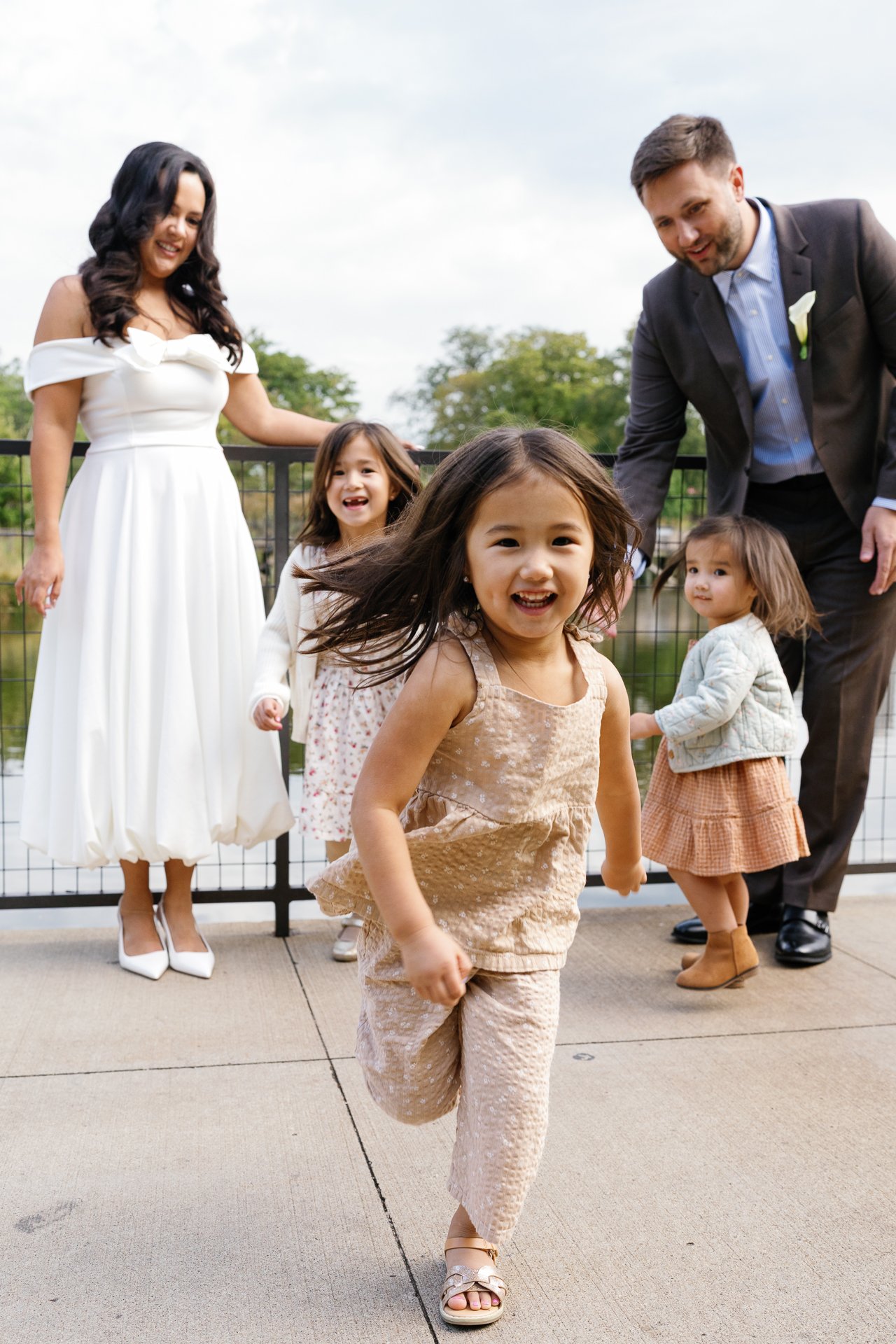 A group of children and two adults enjoy a day outdoors near a pond, with the children running toward the camera and smiling.