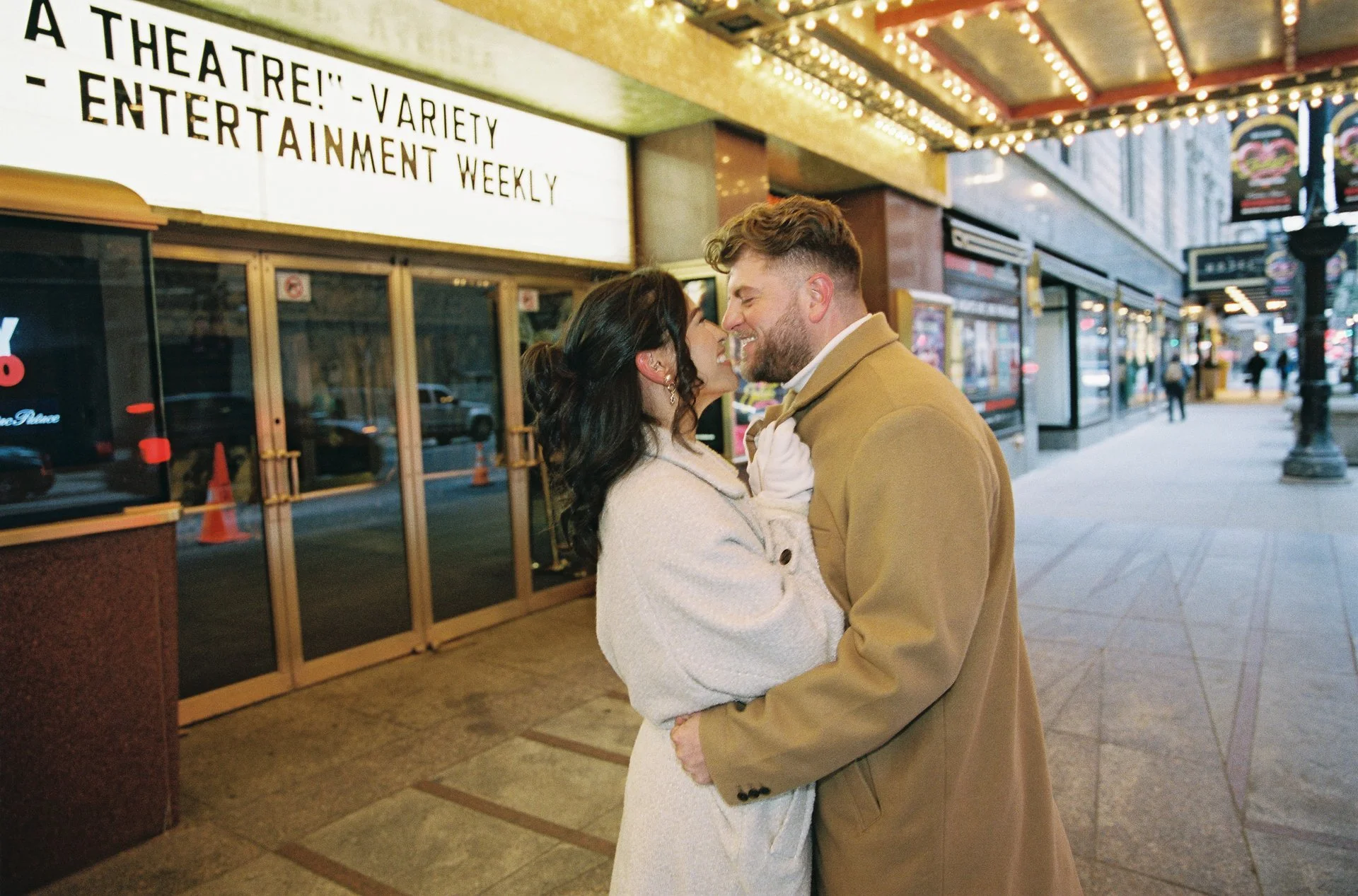 A smiling couple embracing on a city sidewalk at night. The woman has dark hair and wears a light-colored coat, and the man has a beard and wears a tan coat. In the background, there is a theater marquee with bright lights and a sign with black text.