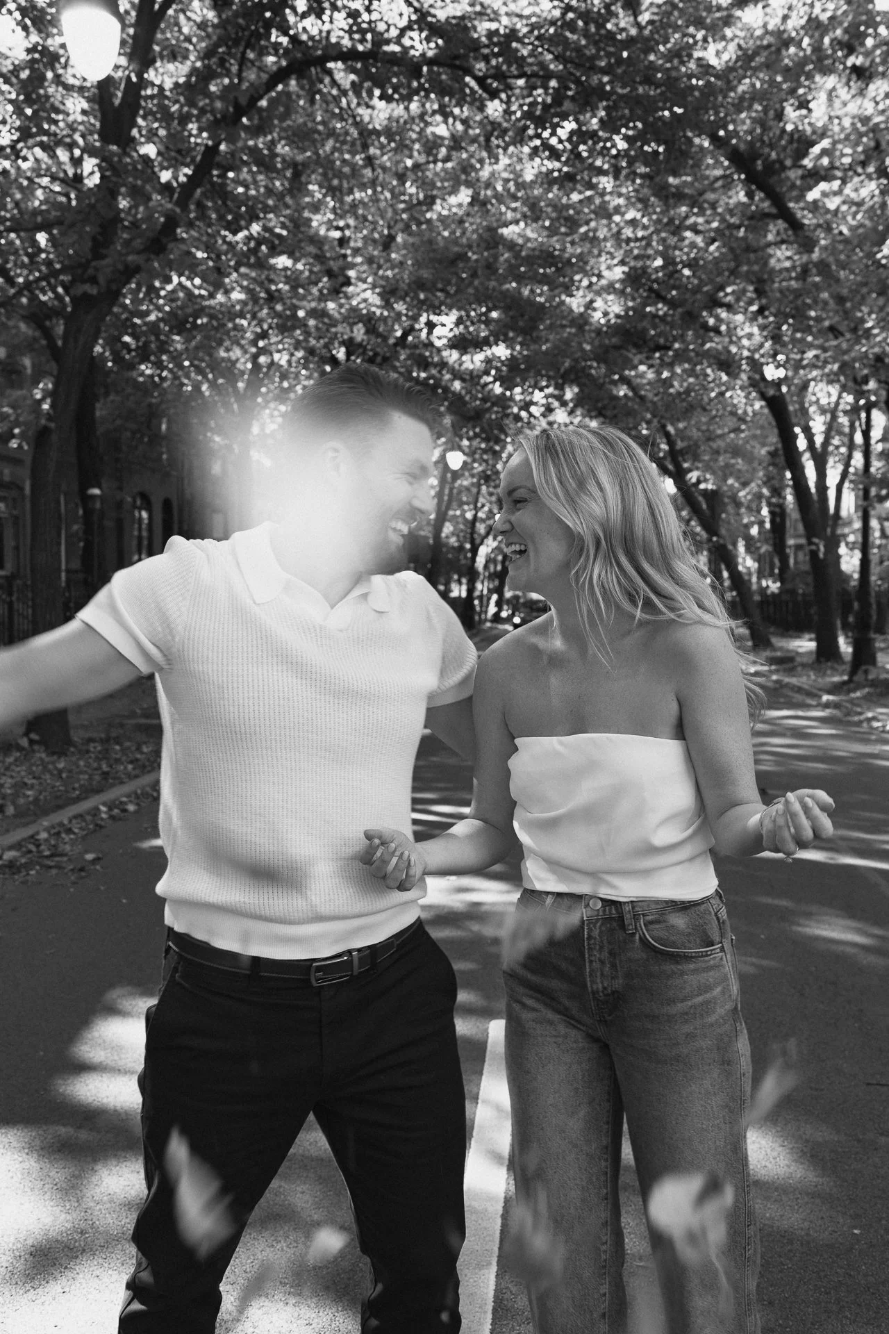 A black-and-white photo of a happy couple laughing and holding hands on a tree-lined street.