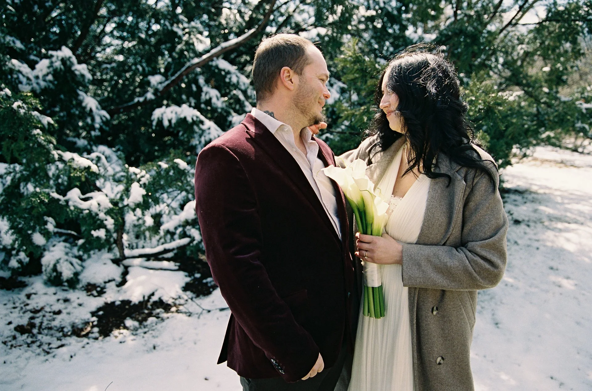 A couple standing close together in a snowy outdoor setting, with trees covered in snow in the background. The woman is holding a bouquet of white calla lilies and smiling at the man.