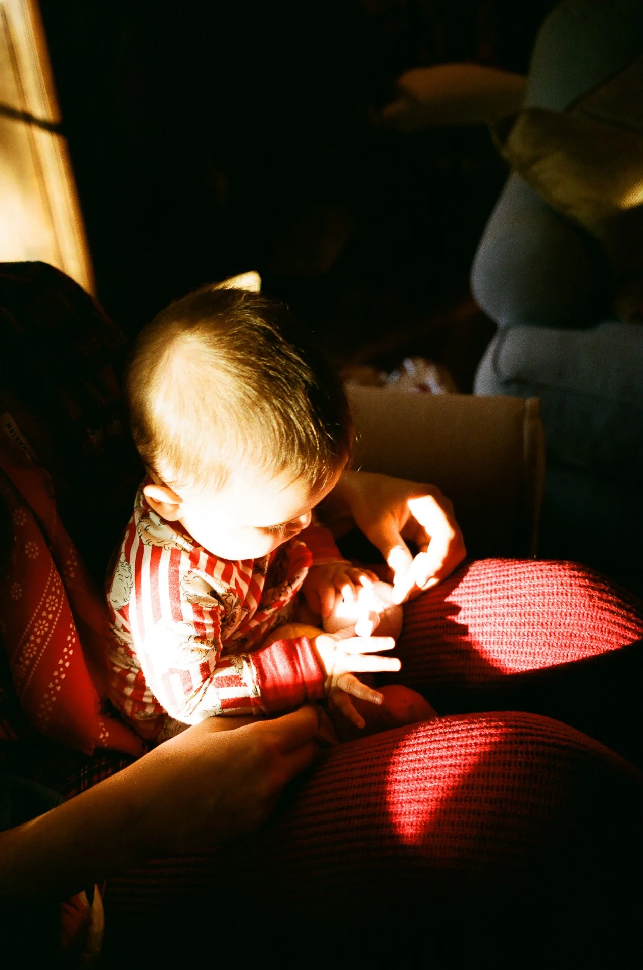 A young child sitting on a person's lap, with sunlight illuminating their face and hands. The child is holding and examining an object, and the scene appears to be indoors with dim surroundings.
