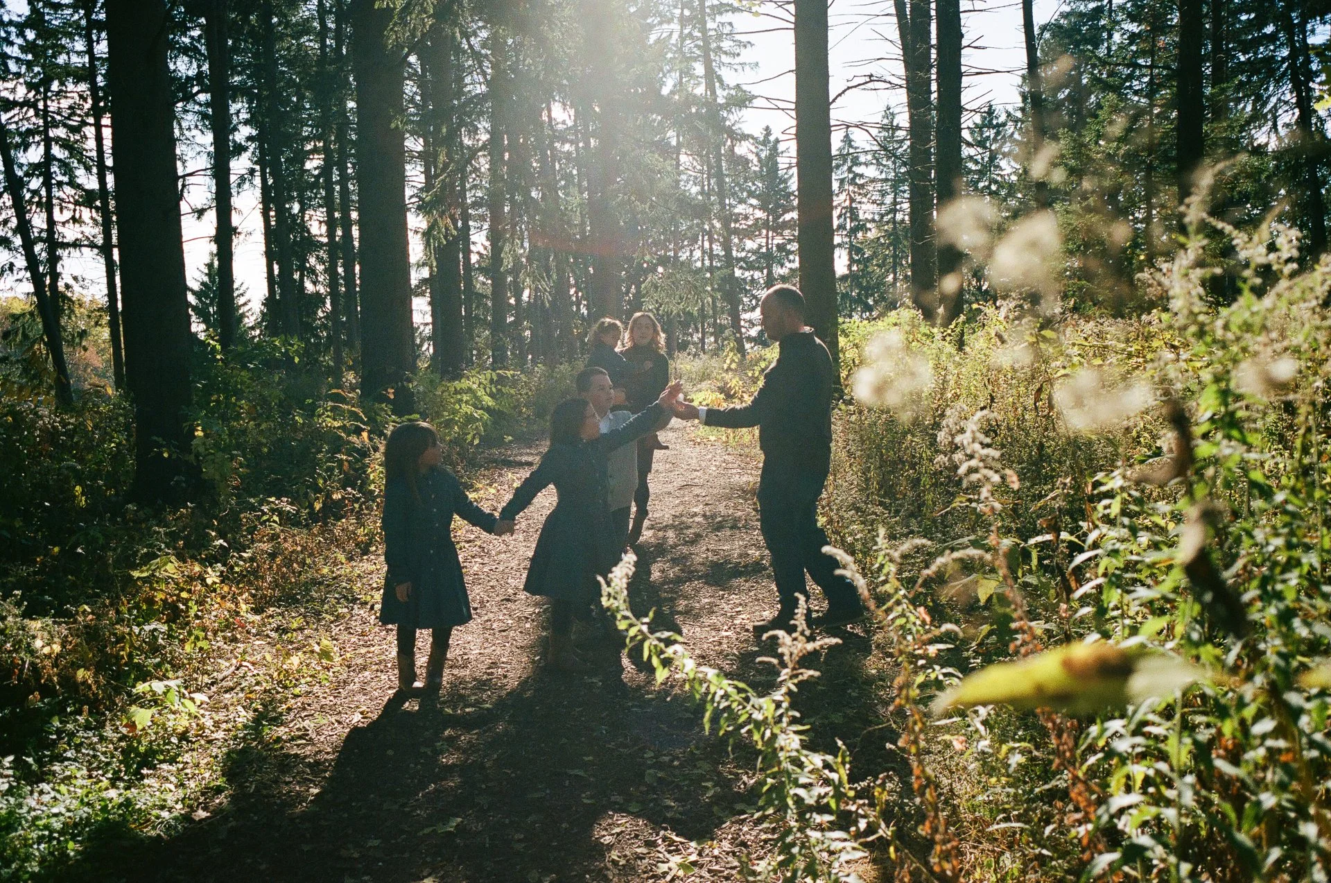 A family of six holding hands and dancing on a forest trail during sunlight.