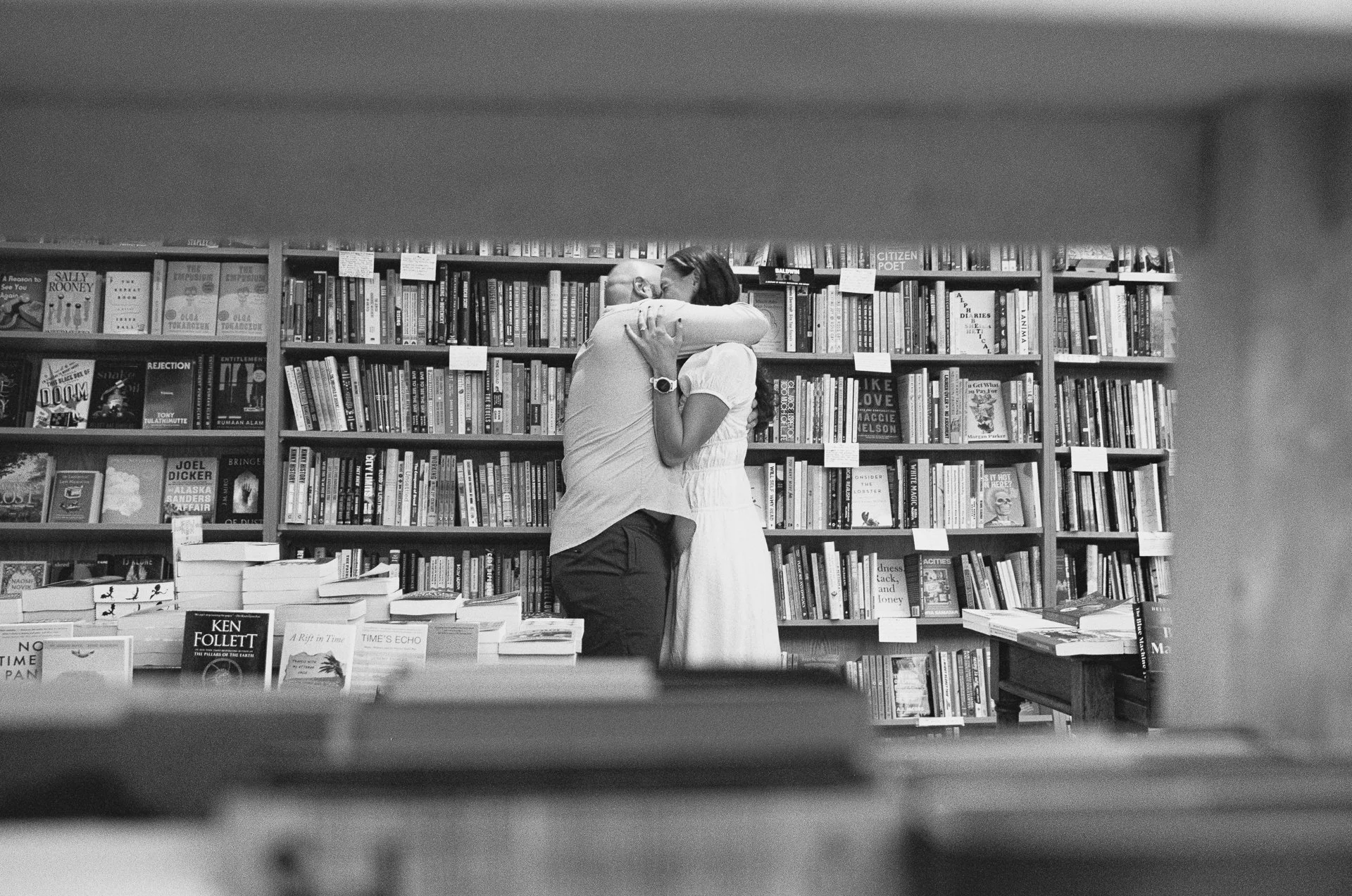 A man and woman embracing inside a bookstore, surrounded by shelves of books.