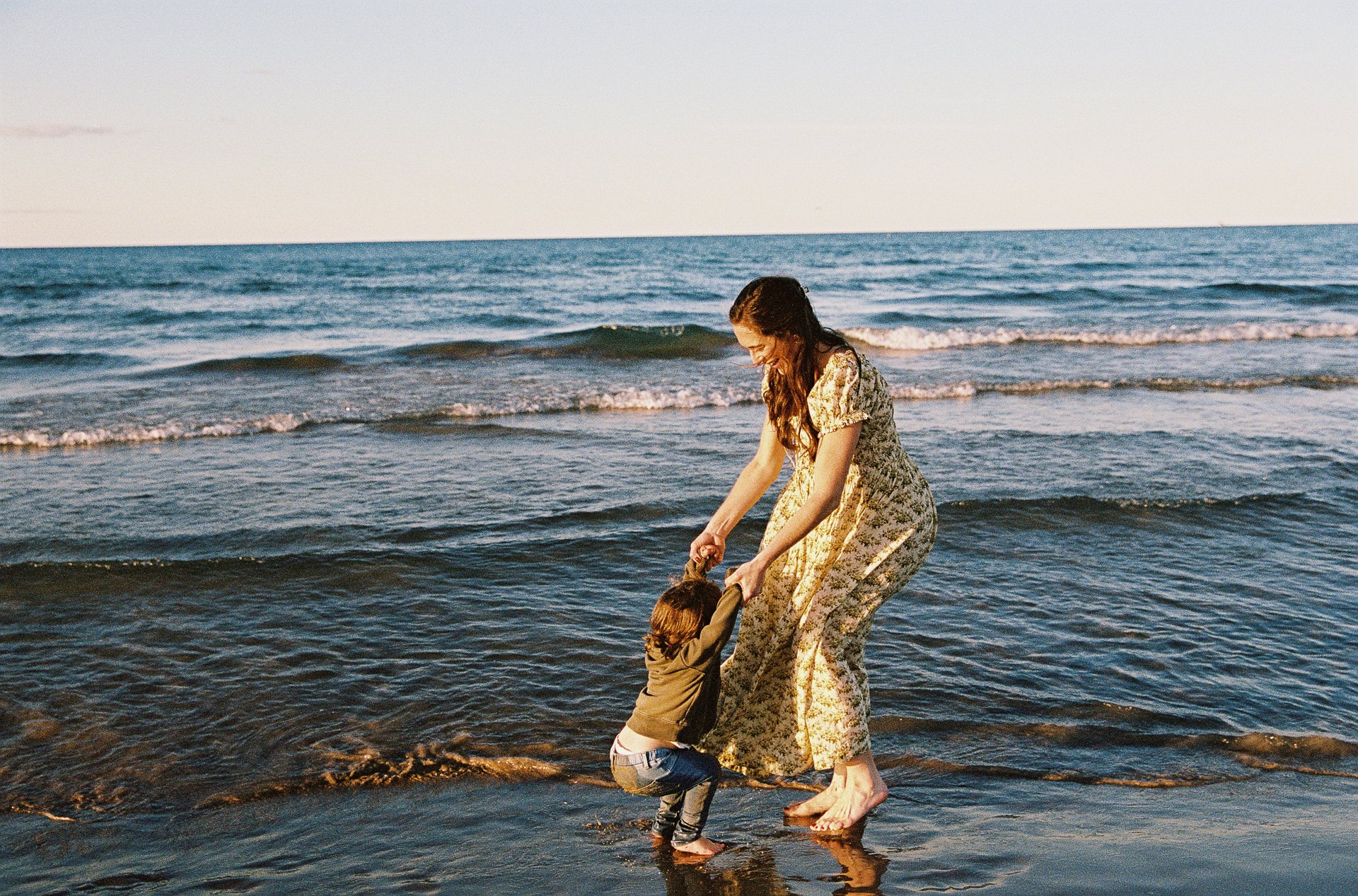 A woman and a young child playing in the shallow surf at the beach during sunset.