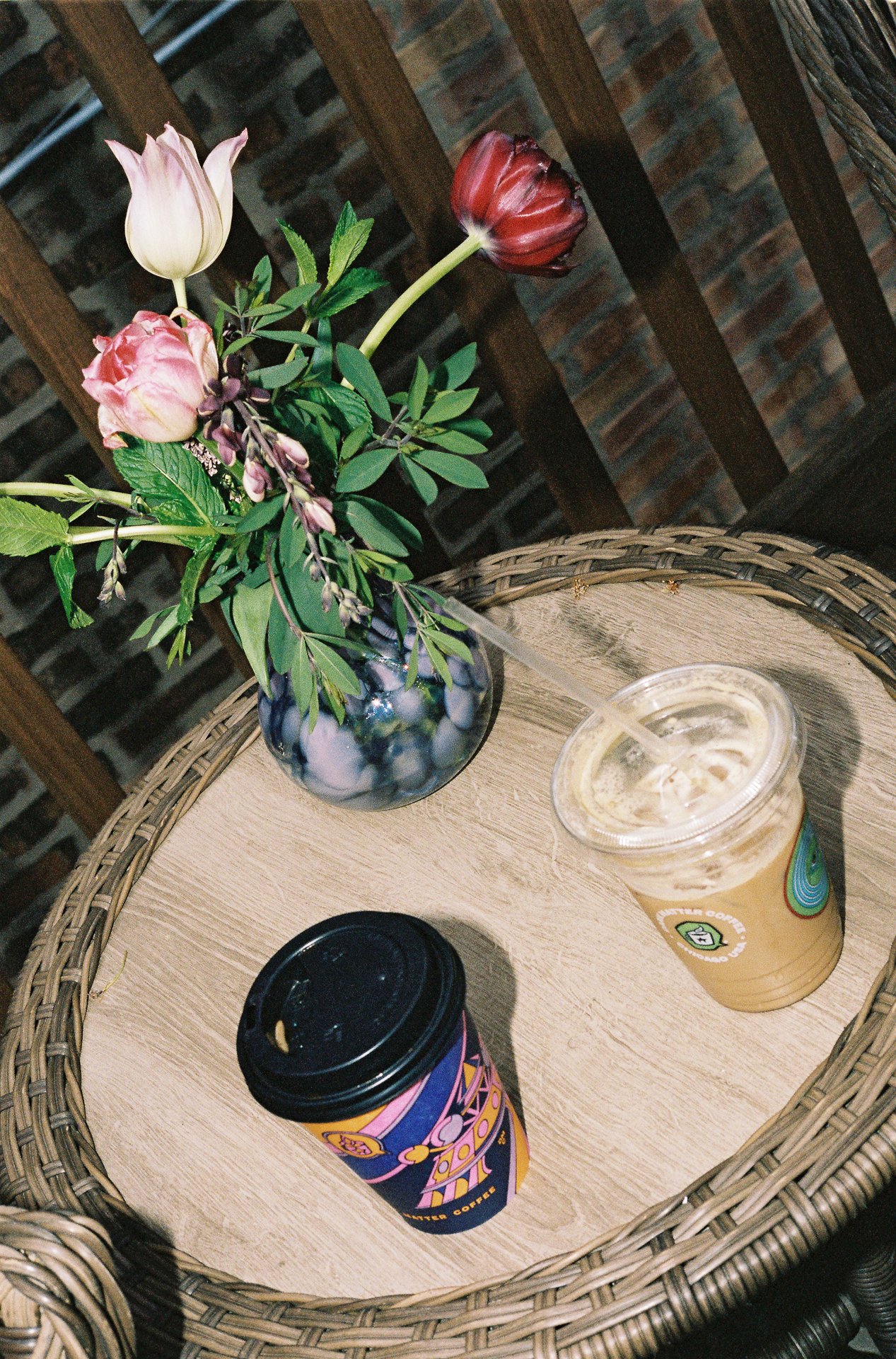 A small round table with a wicker edge, holding a vase of mixed flowers and two coffee cups, one black with a pink and yellow design, the other clear with a light-colored drink inside.