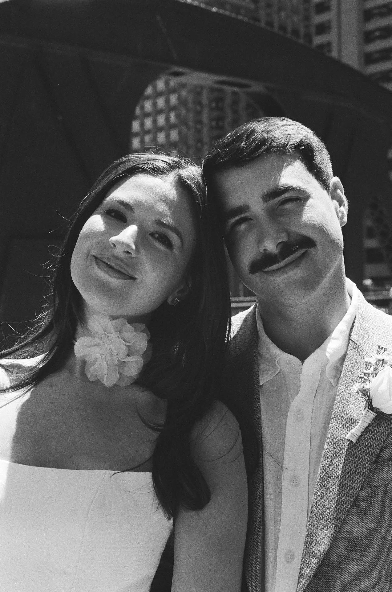 A black and white photo of a smiling couple taking a selfie outdoors in an urban setting, with tall buildings in the background.