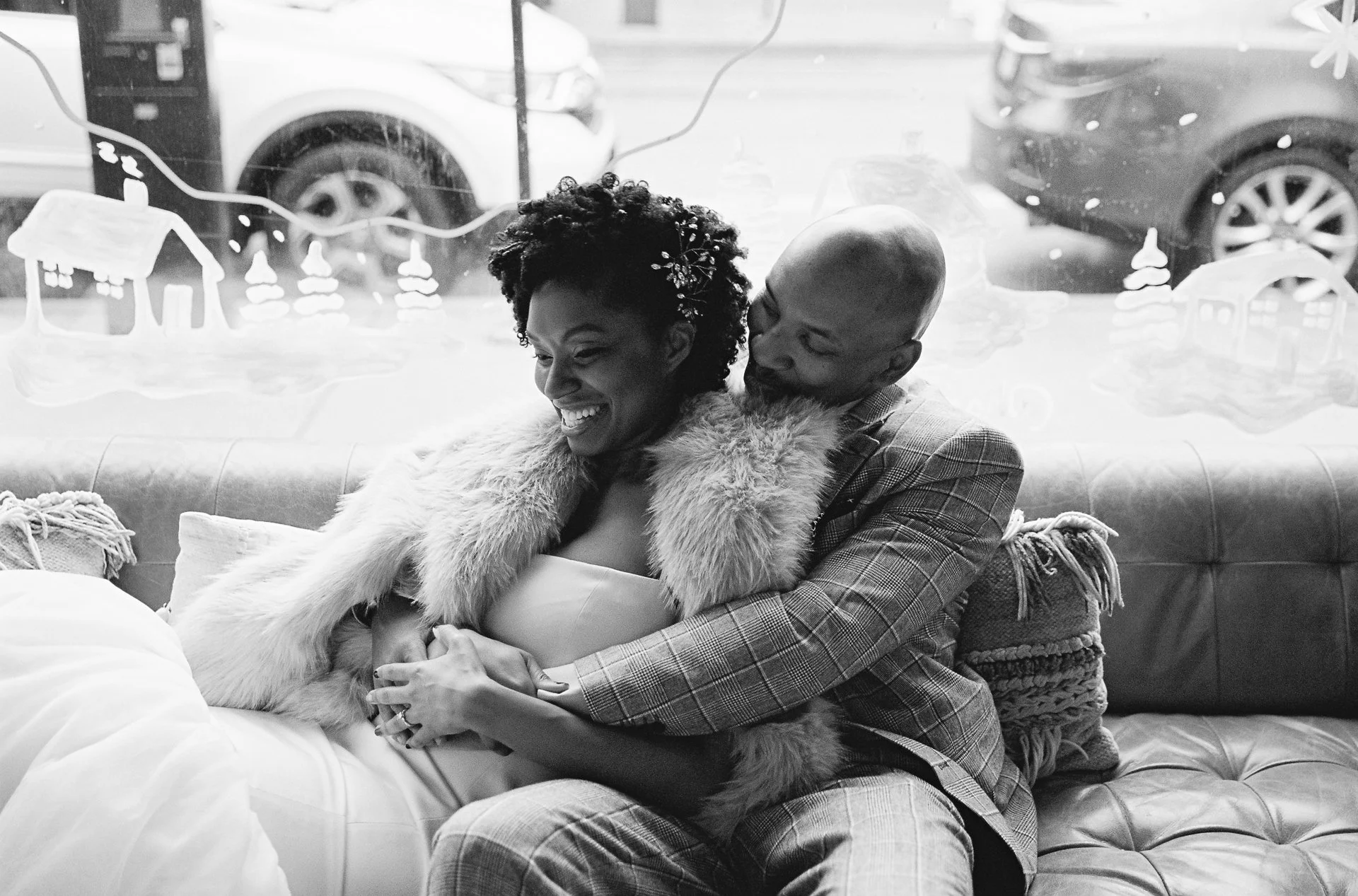 A joyful couple sitting on a couch, with the woman in a wedding dress and fur stole, and the man in plaid suit, smiling and embracing each other, with holiday window decorations in the background.