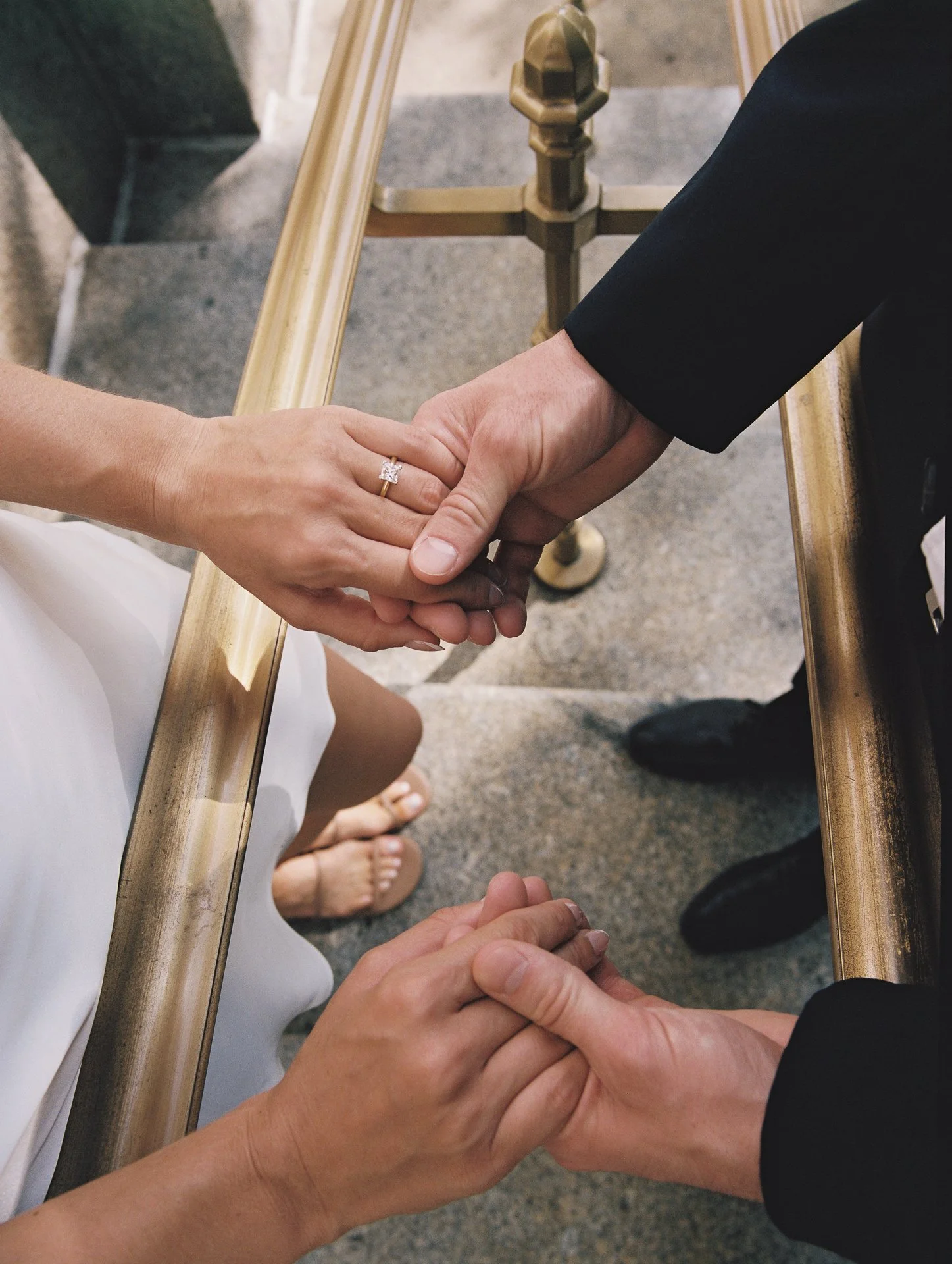 A bride and groom are holding hands during their wedding ceremony, with the bride wearing a white dress and an engagement ring, and the groom wearing a black suit.