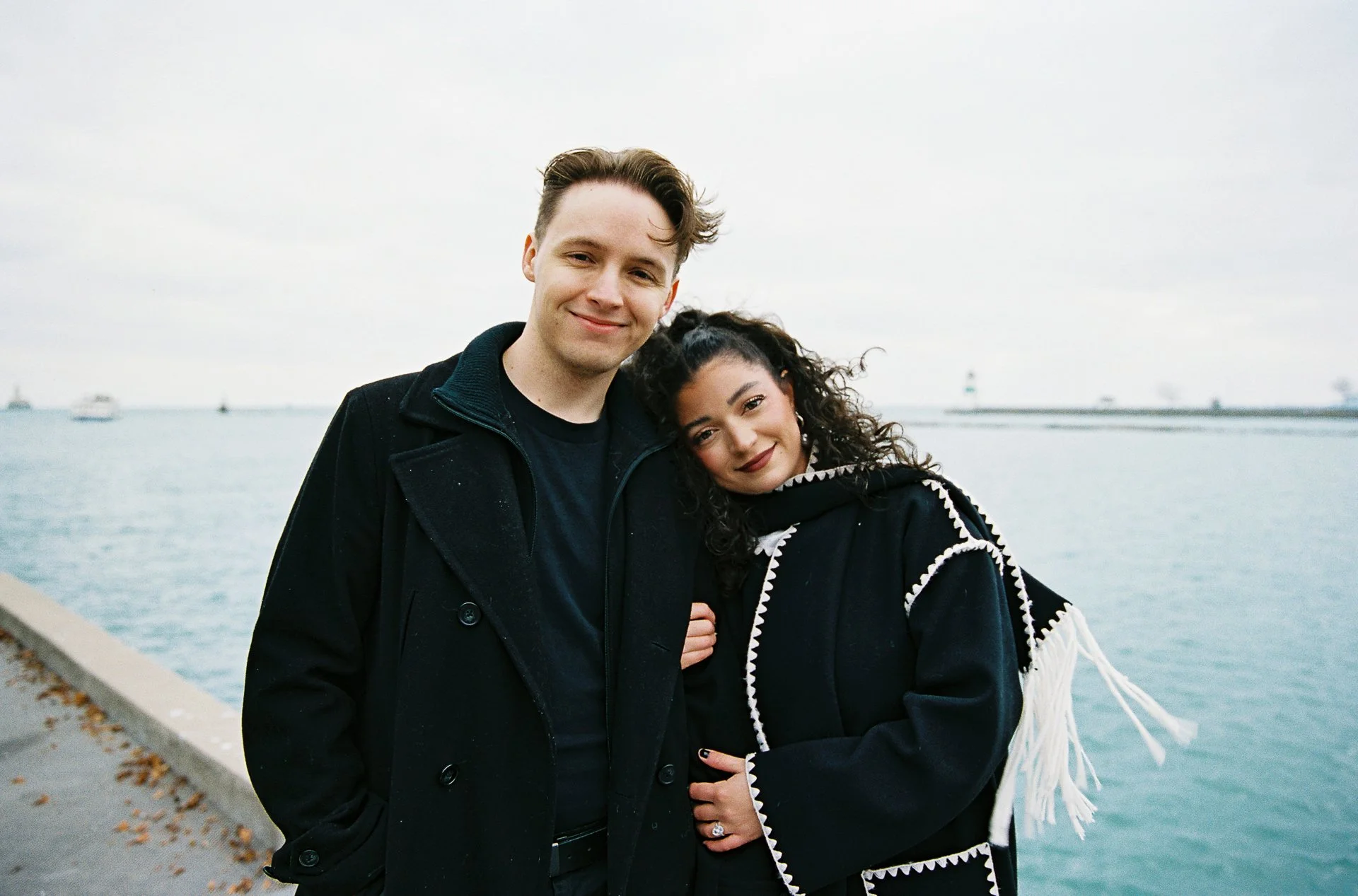 A young couple stands near a waterfront, smiling and cuddling under an overcast sky with boats in the distance.