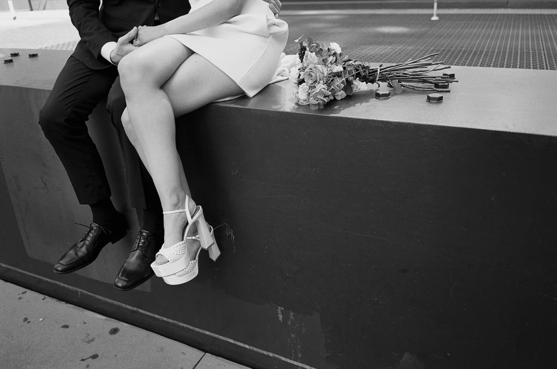 A couple sitting on a ledge, with a bouquet of flowers beside them, in black and white.