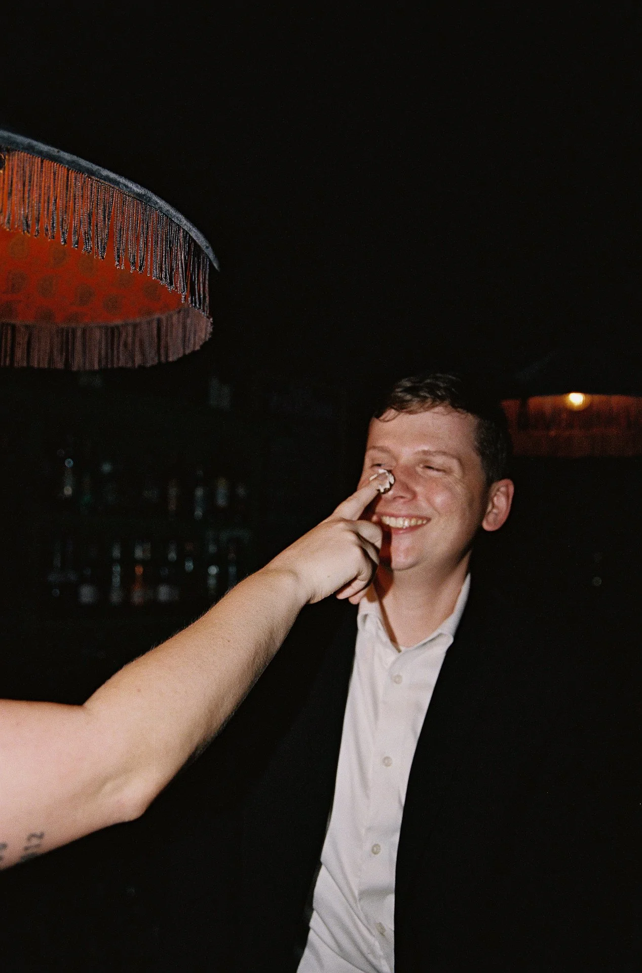 A young man in a suit smiling as someone playfully touches his nose, in a dimly lit room with a red lampshade.