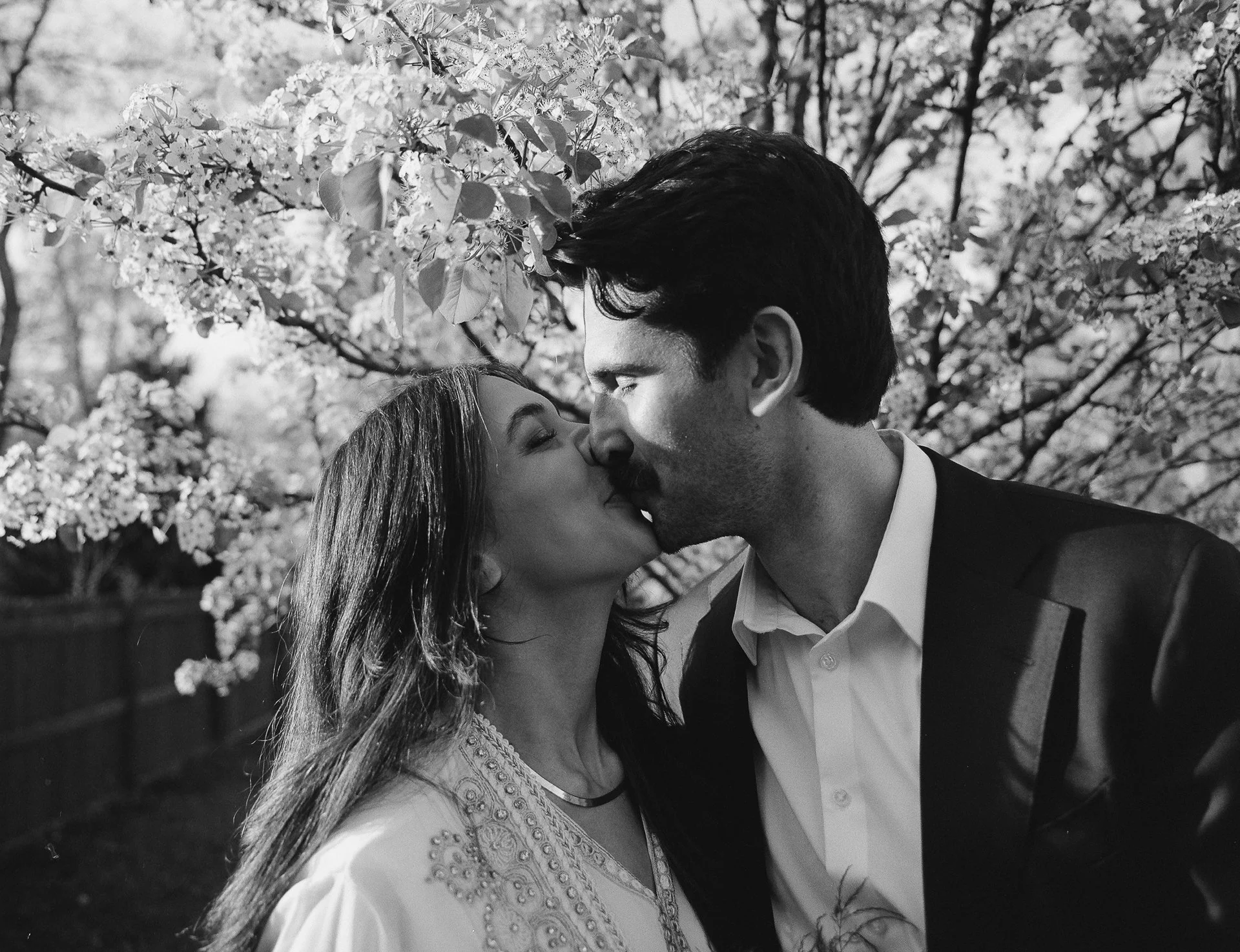 A black-and-white photo of a couple kissing outdoors, surrounded by flowering trees.