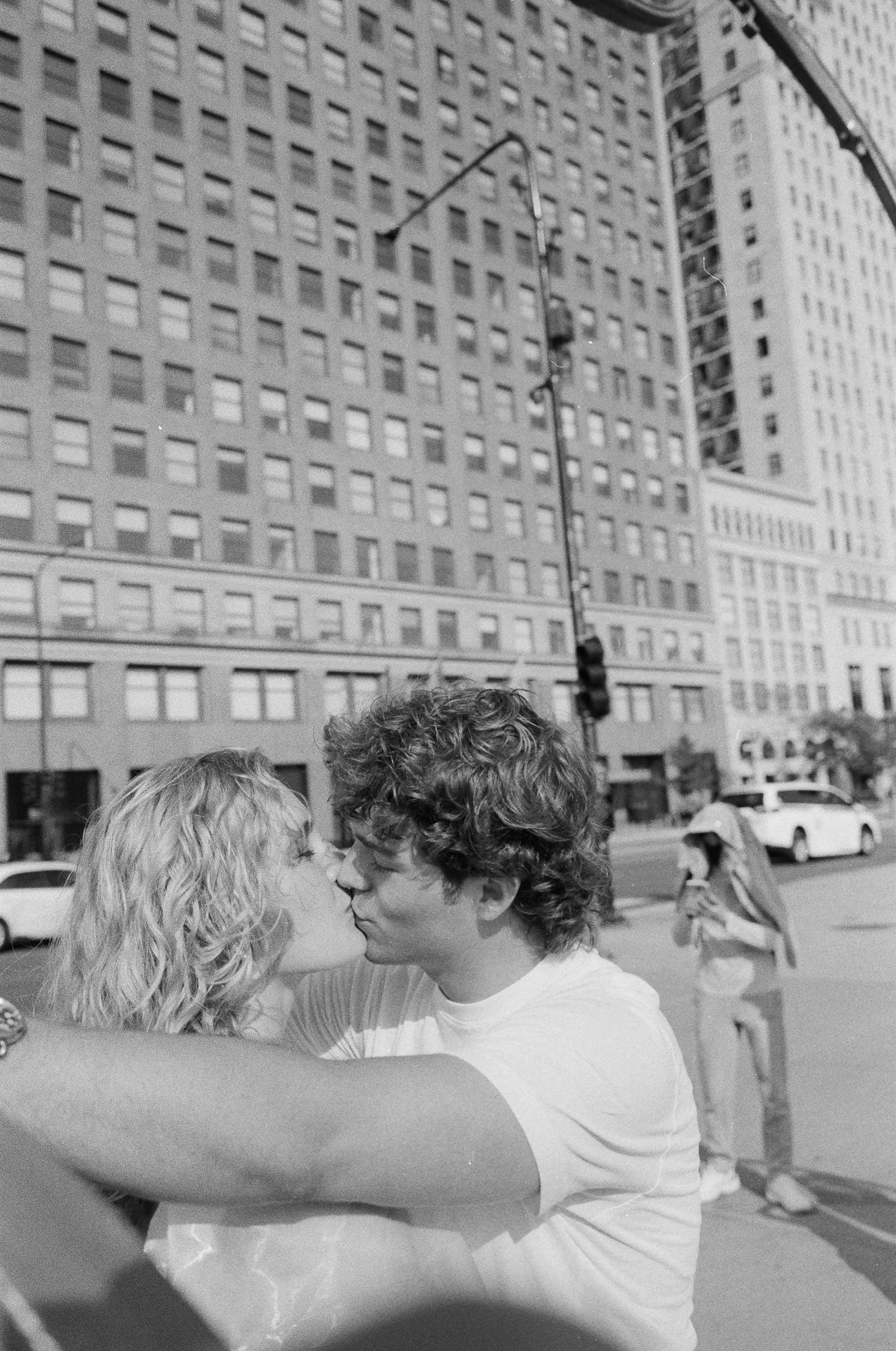 A black-and-white photo of a young couple with curly hair sharing a kiss on a city street, with a tall building and a person walking in the background.