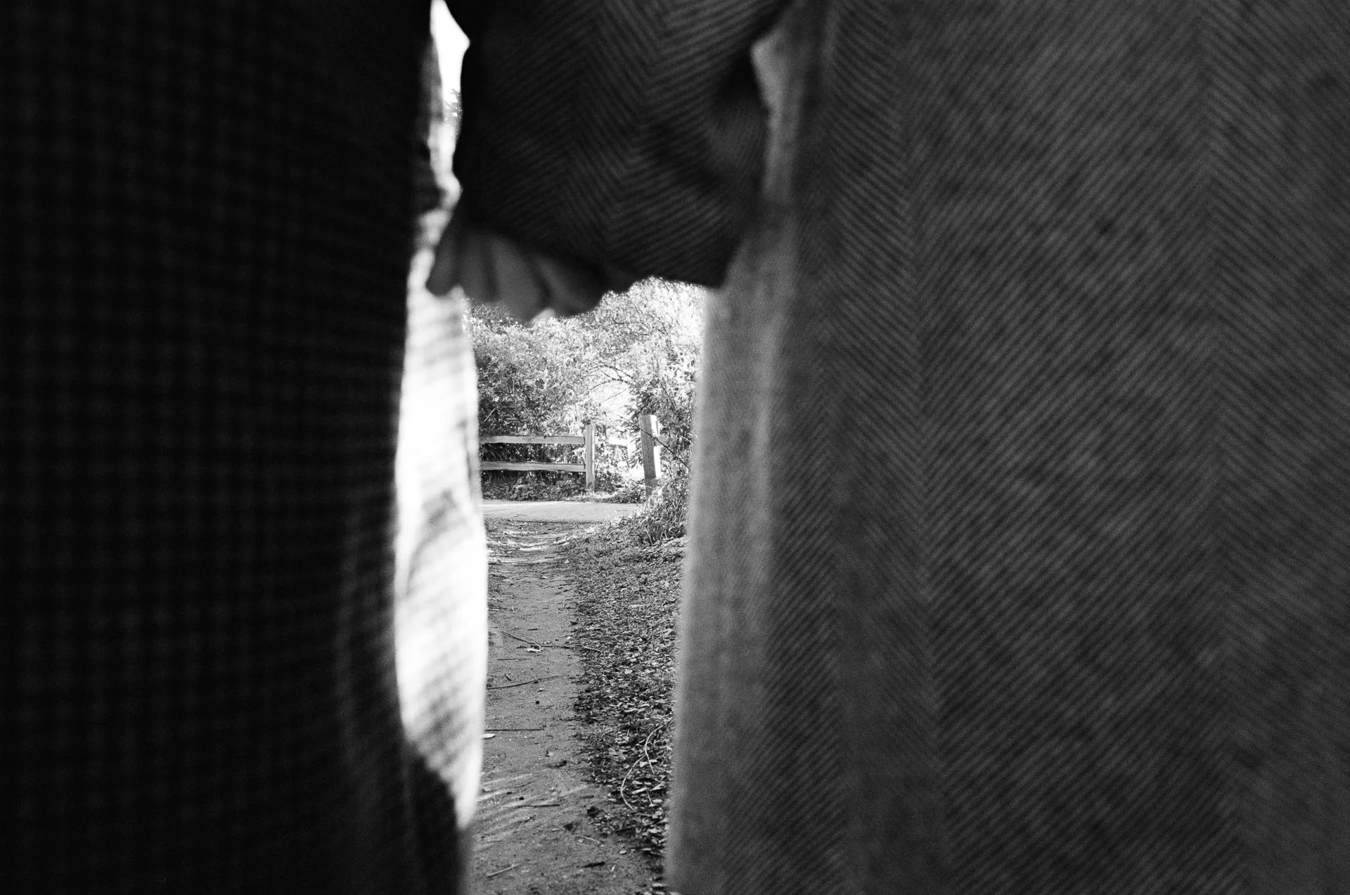 A black-and-white photo taken through a narrow gap between two bundles of fabric or clothing, showing a dirt path, trees, and a wooden fence in the background.