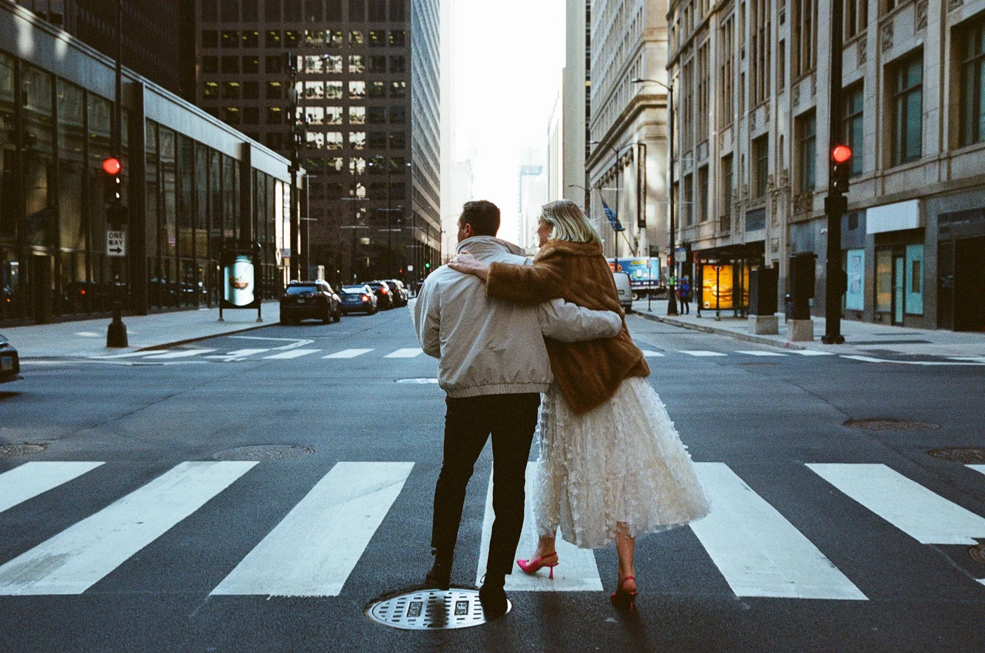 A couple walking arm in arm across a city street crosswalk in a downtown area with tall buildings and traffic.
