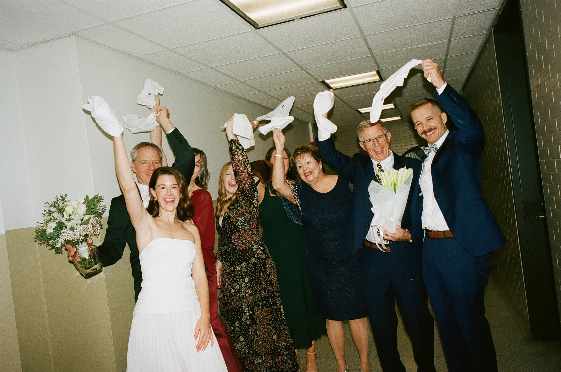 Group of people celebrating a wedding, some holding paper towels in the air, smiling and joyfully interacting in a hallway.