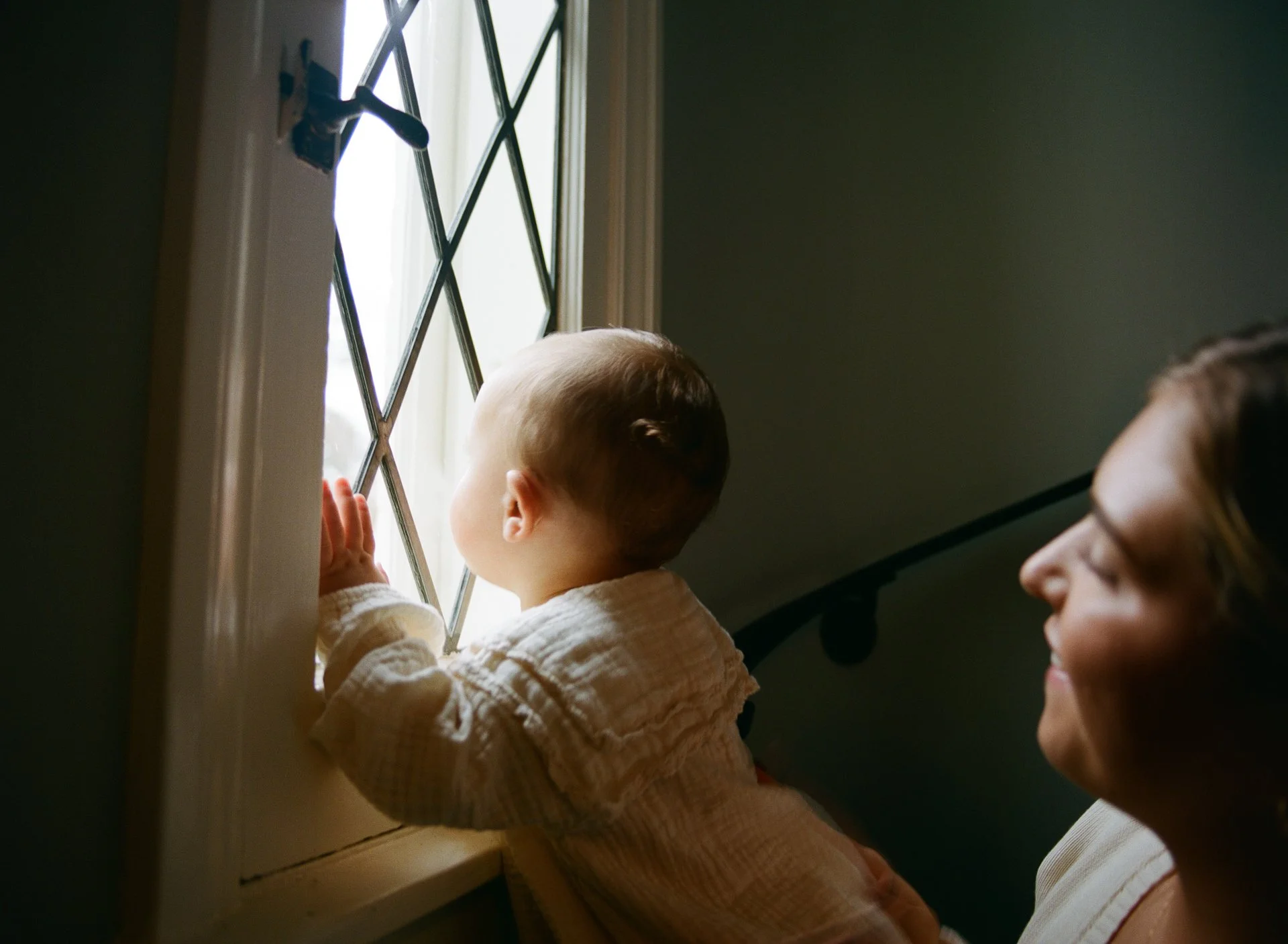 A young child looking out a window with a woman smiling behind them.