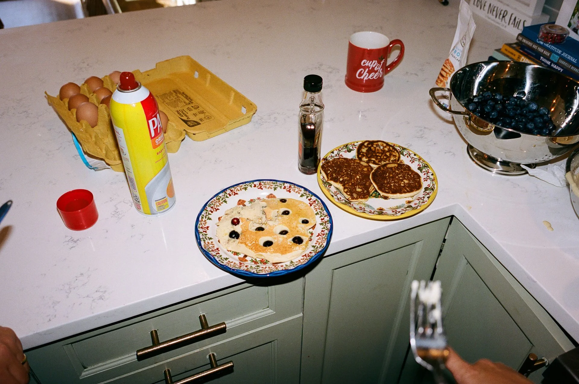 A kitchen counter with three plates of pancakes topped with berries, a carton of eggs, a bottle of syrup, a mug with 'cup of Cheer' written on it, a bowl of blueberries, and other breakfast items.