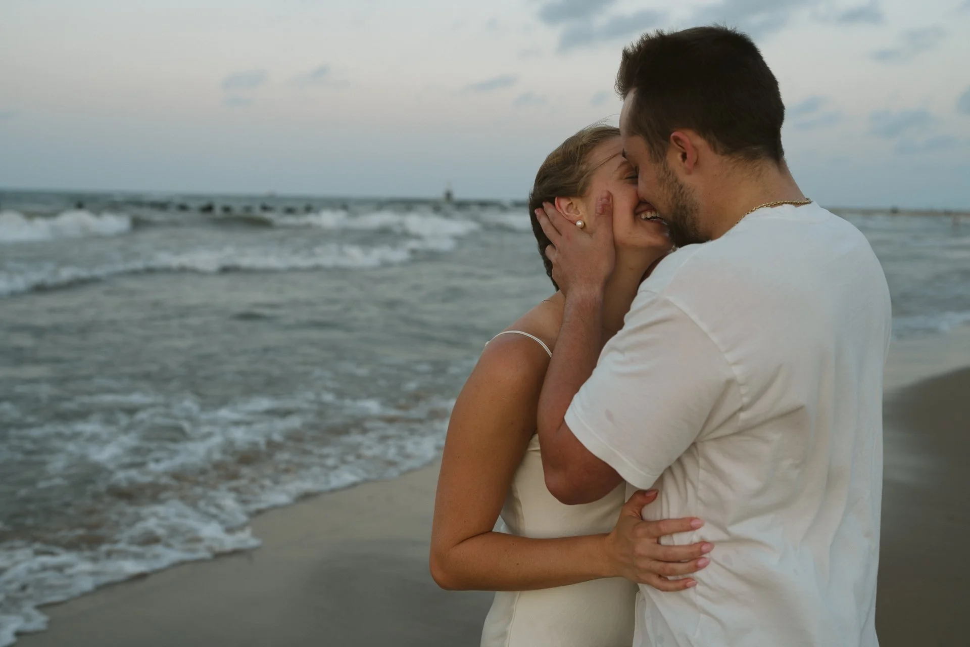 A couple embracing and smiling on the beach near the shoreline with waves in the background.