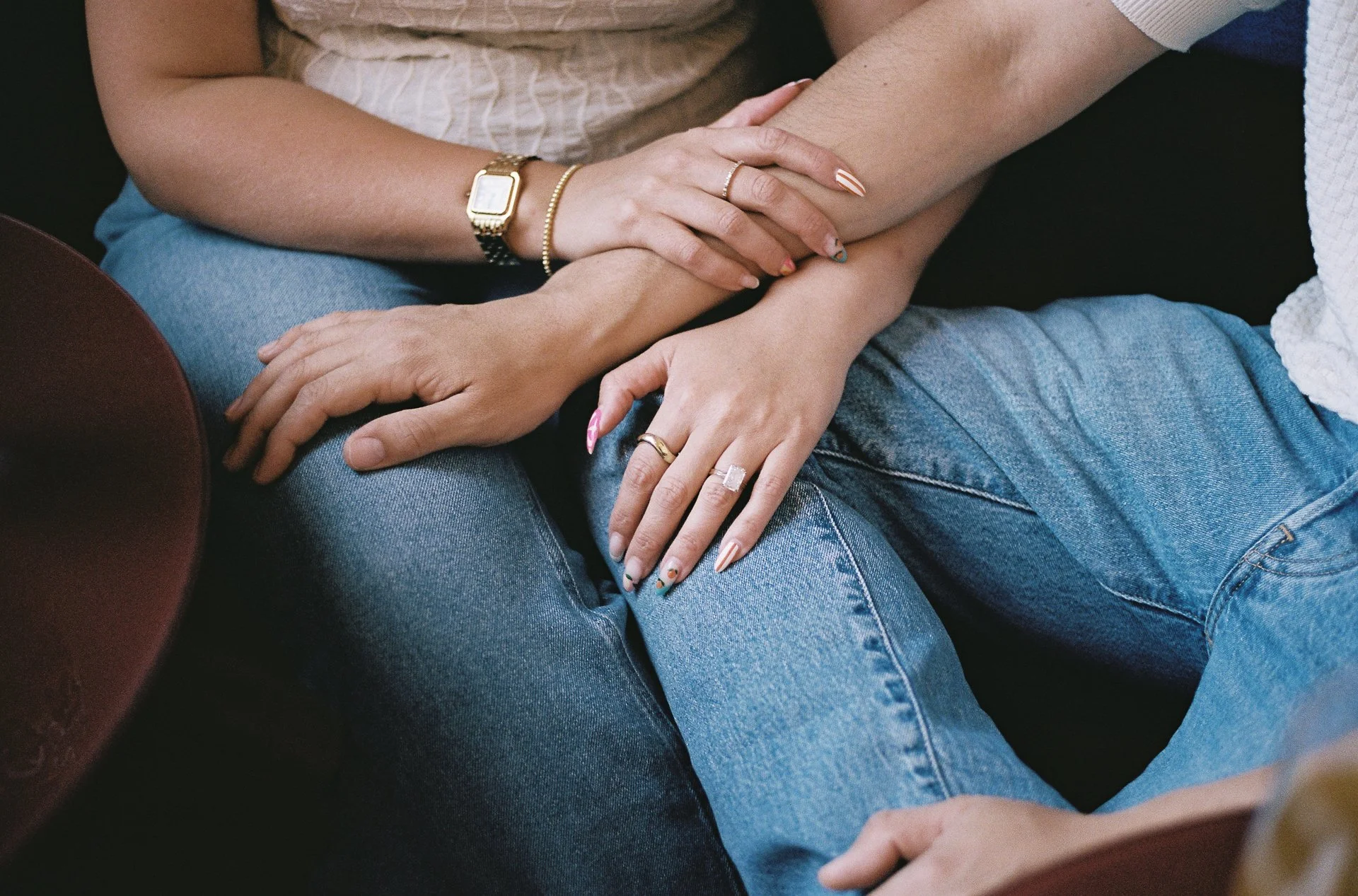 Close-up of two people sitting together, holding hands with interlocked fingers, wearing casual jeans. One person wears a gold watch and jewelry, the other has a large ring with a prominent gemstone.