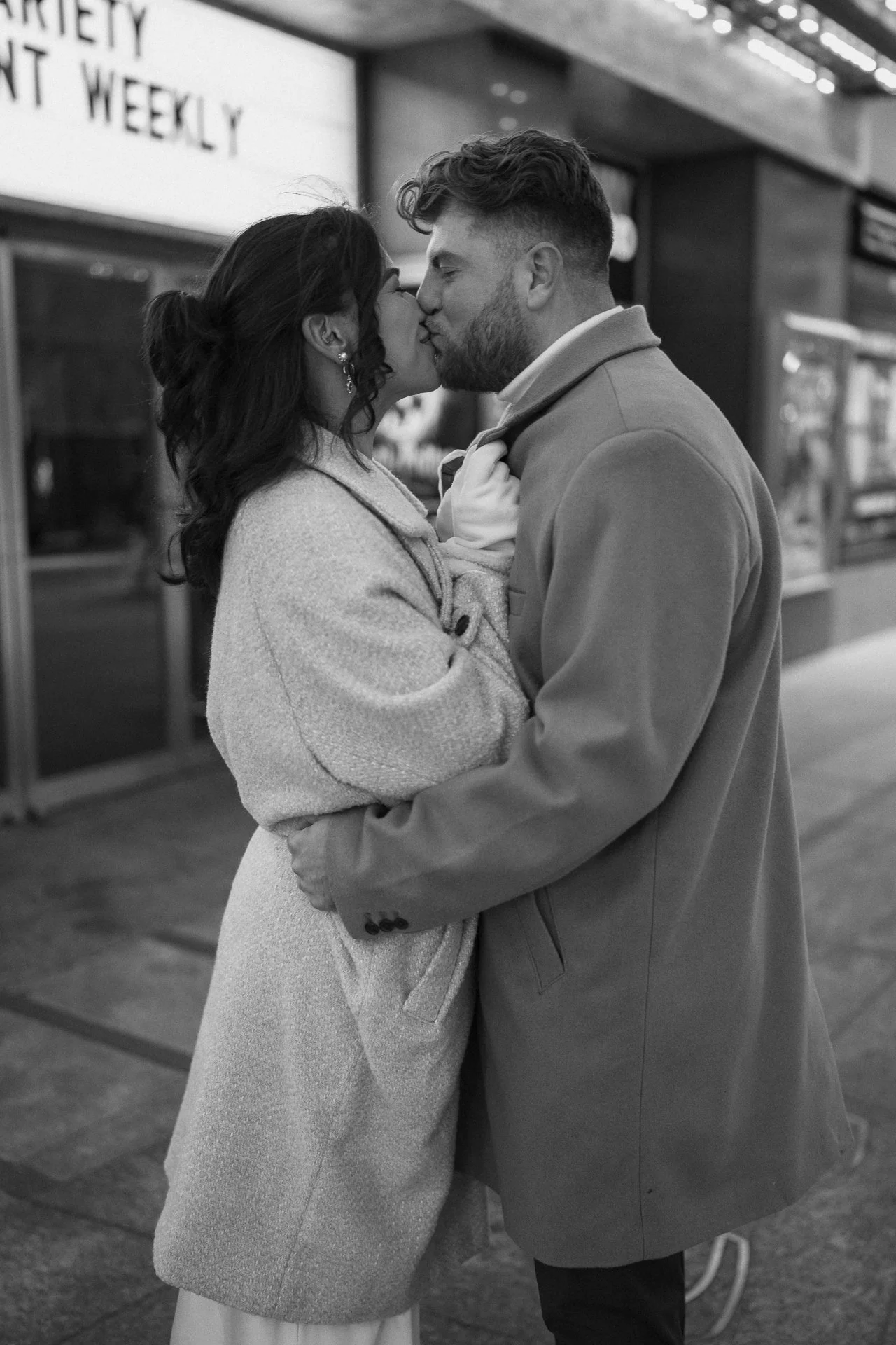 A man and woman sharing a kiss on a city street at night, dressed warmly, with a store sign visible in the background.