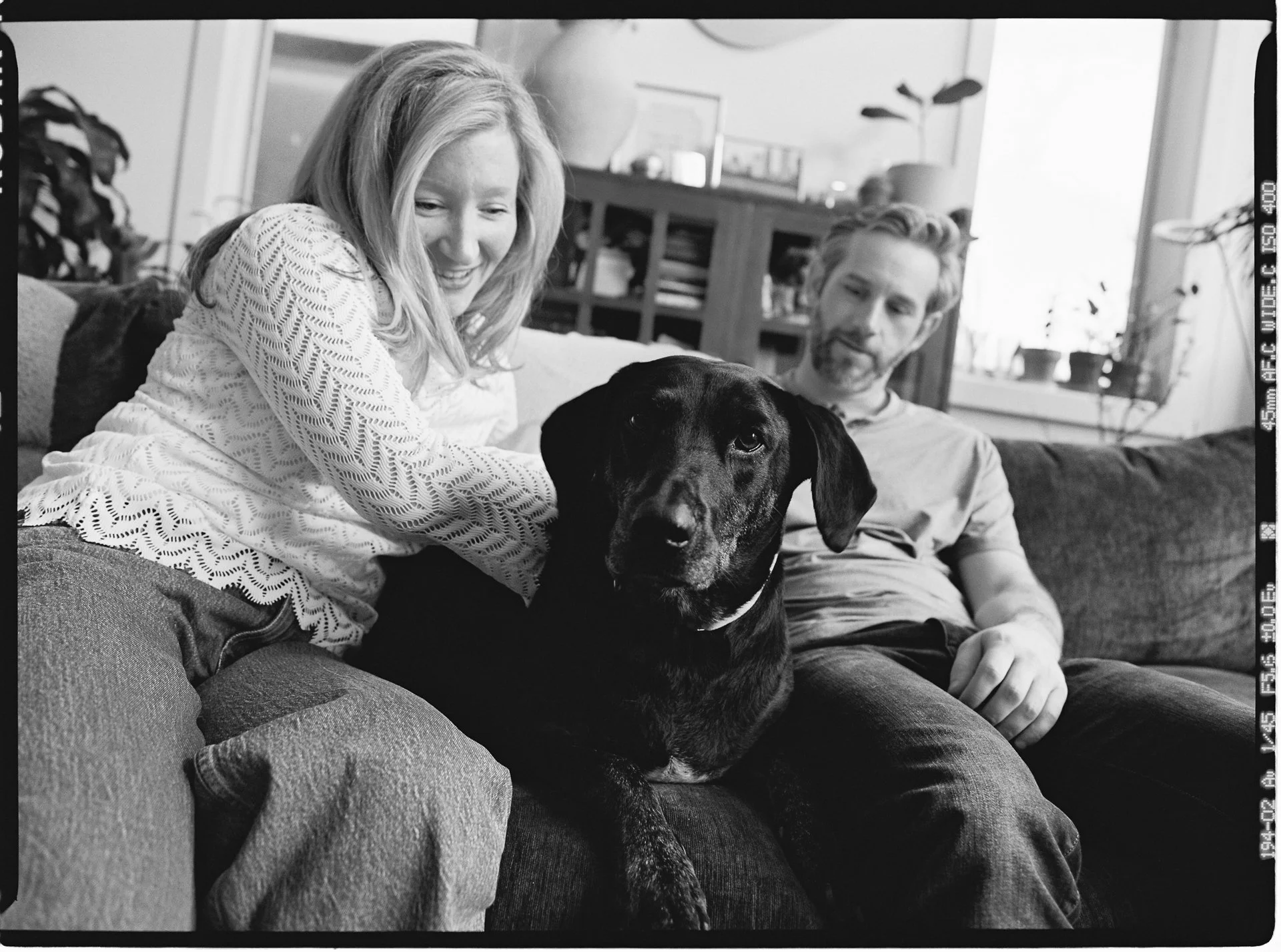 A woman, a man, and a black dog sitting on a couch in a living room, smiling and looking at each other.
