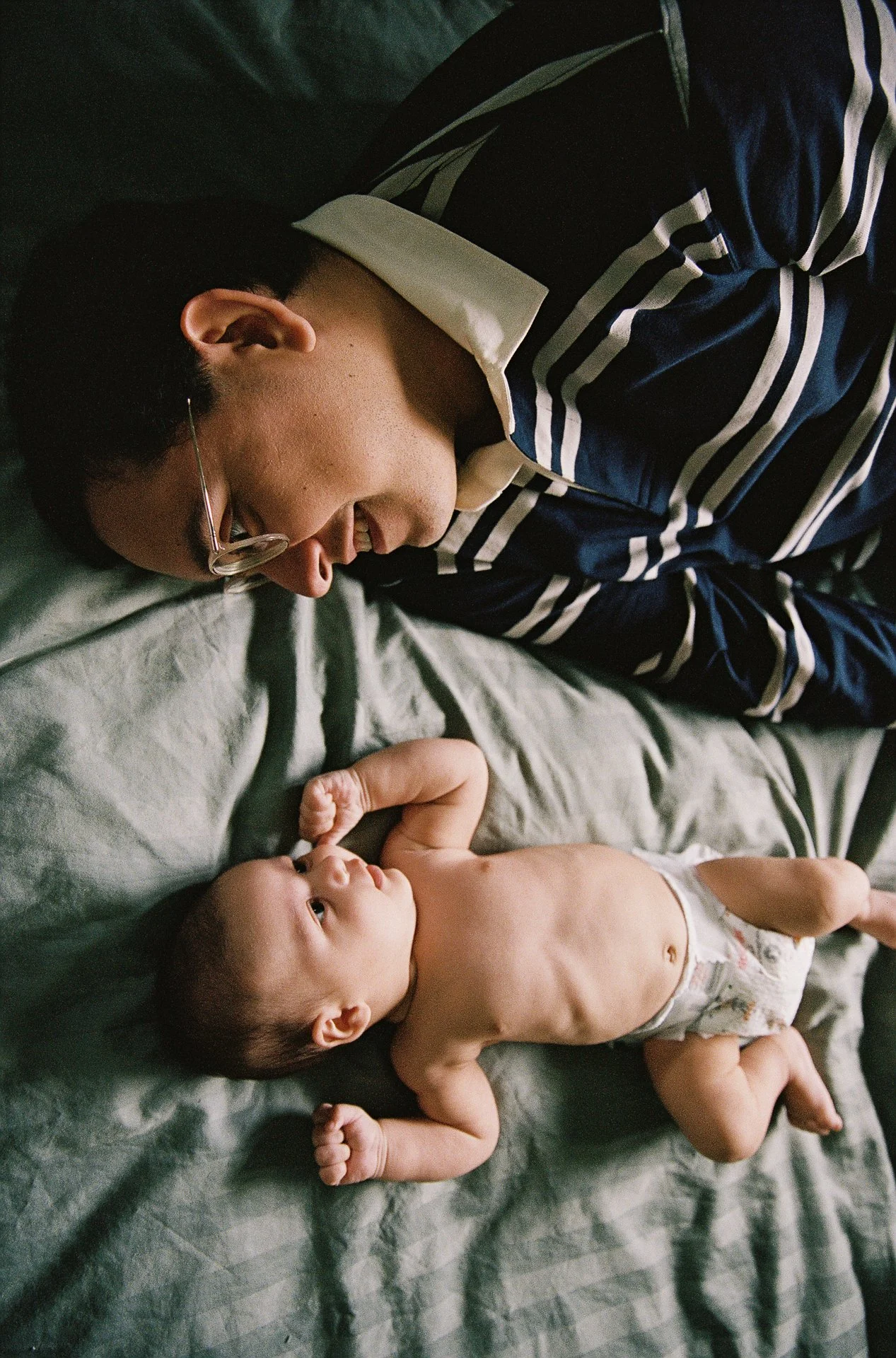A man lying on a bed next to a baby, both looking at each other. The man is wearing a striped shirt and glasses, smiling at the baby who is lying on the bed in a diaper.