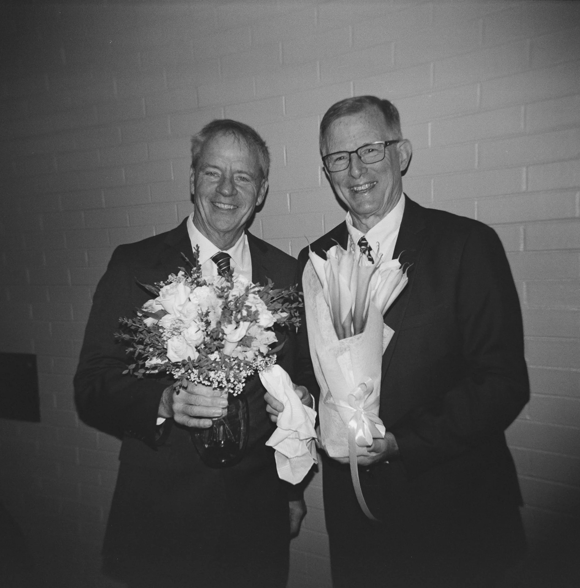 Two elderly men in suits holding bouquets of flowers, smiling at the camera, standing against a brick wall.