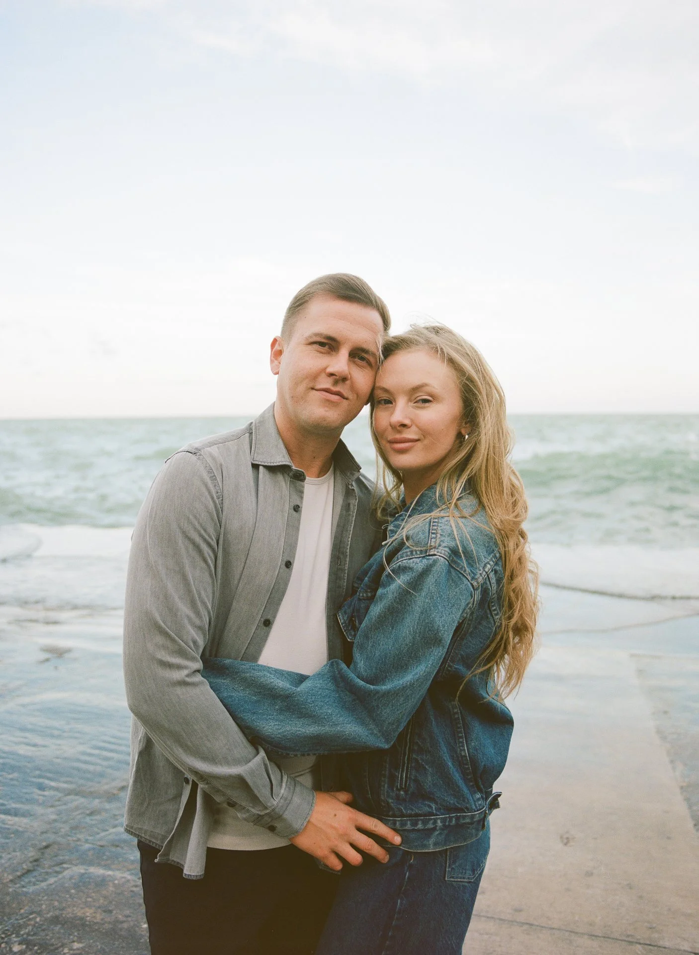 A young couple standing close together on a beach, with the ocean and a cloudy sky in the background.