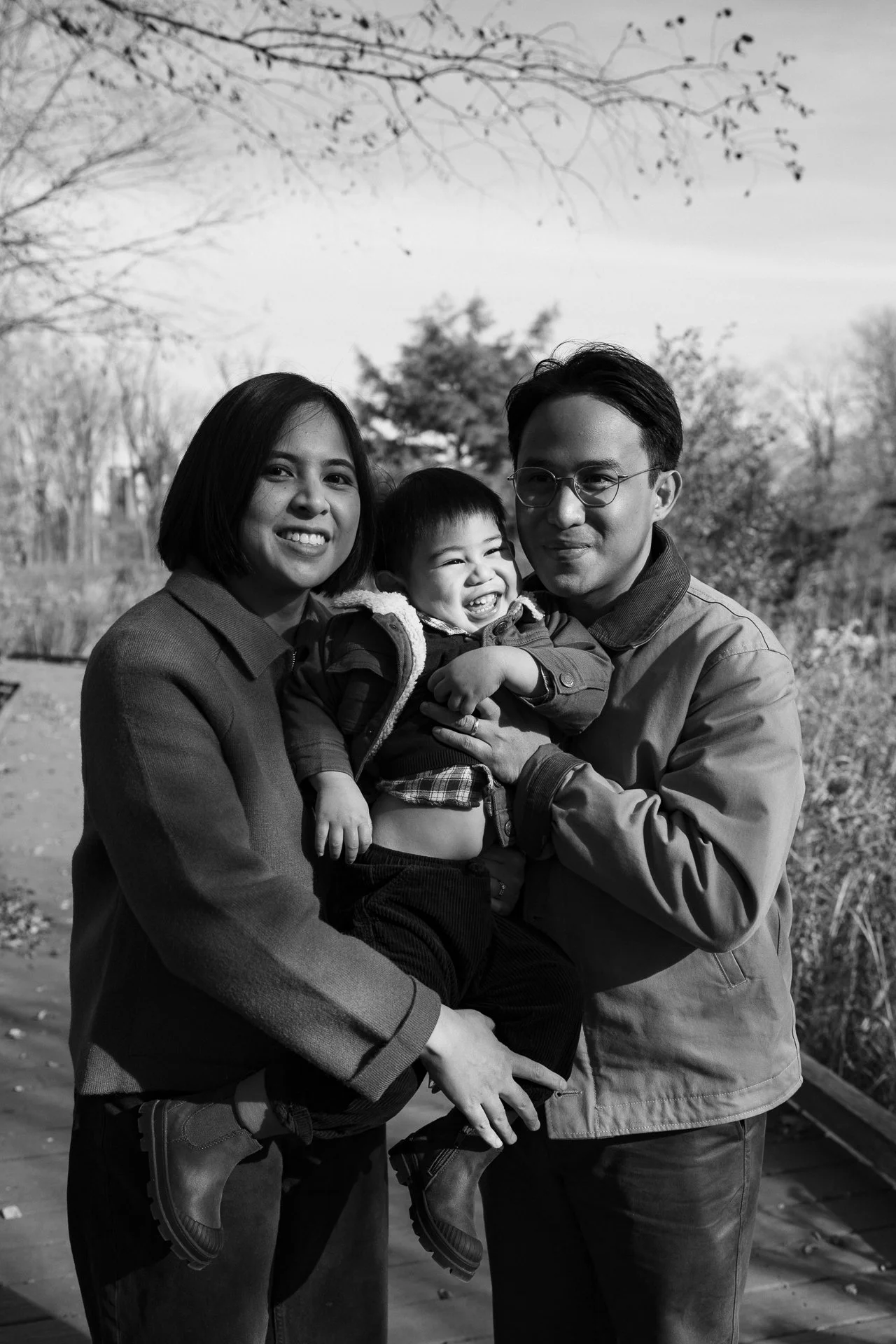 A family of three outdoors during autumn, with the mother and father holding their laughing young son between them. The background features trees with few leaves and a cloudy sky.