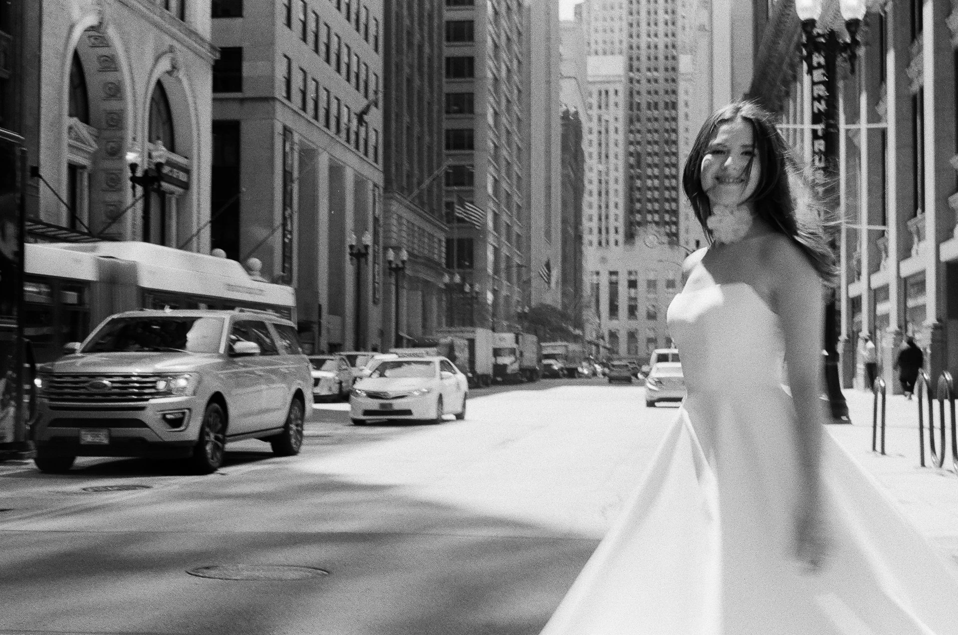 A woman in a white dress standing on a city street with tall buildings and cars.