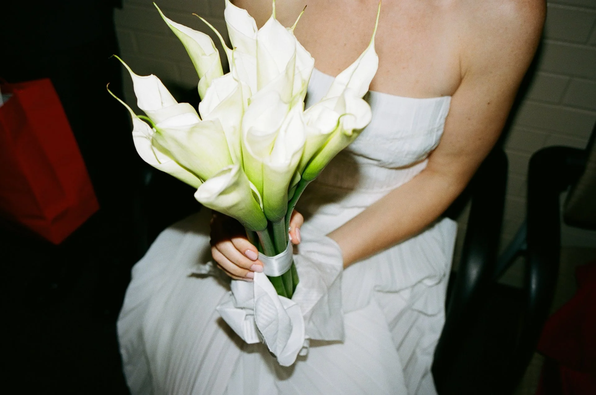 A woman in a white strapless dress holding a bouquet of white calla lilies with green stems, wrapped in white ribbon.