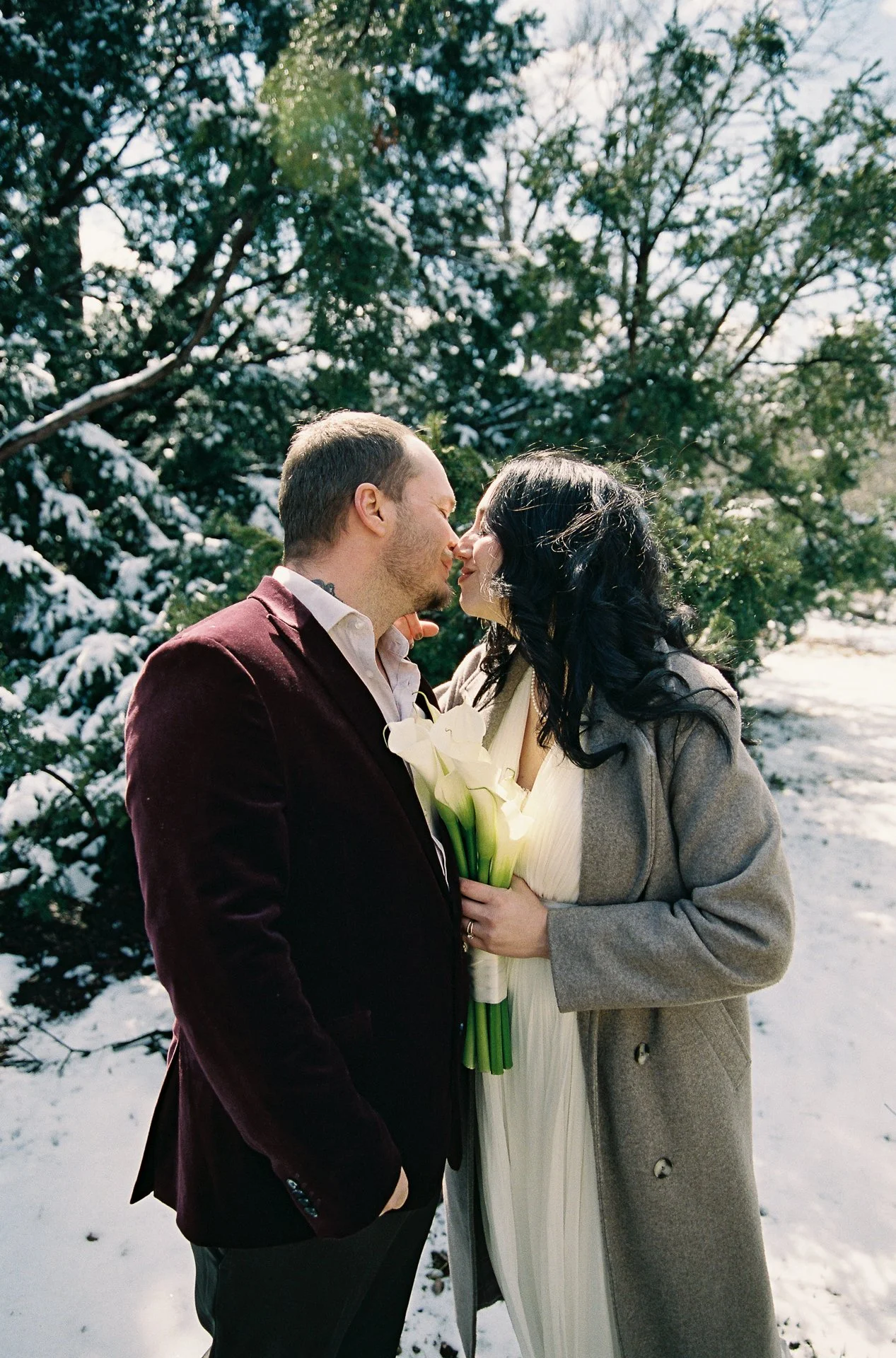 A couple is kissing outdoors in a snowy landscape, holding a bouquet of white calla lilies, with evergreen trees in the background.