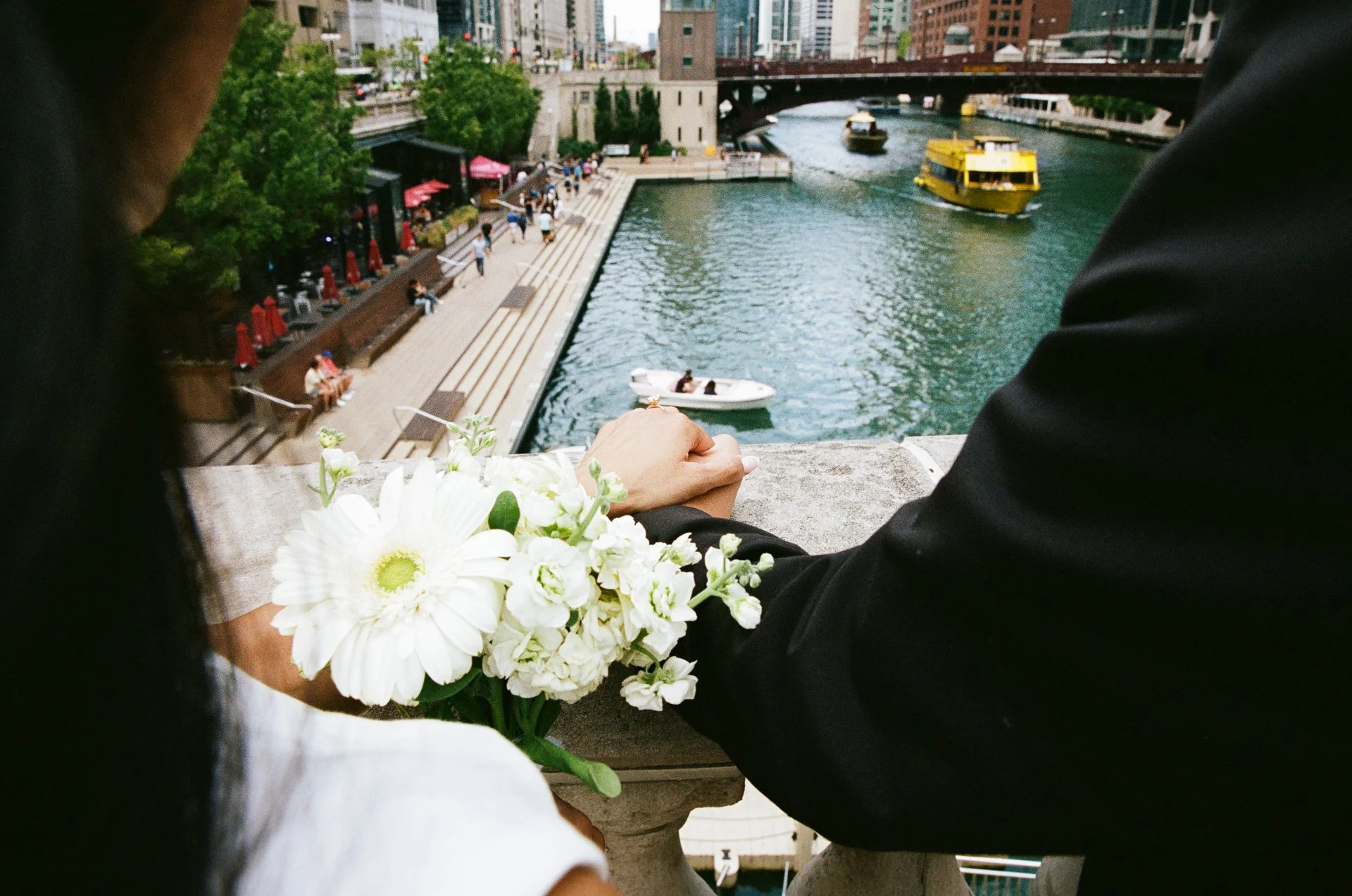 A couple holding hands leaning over a bridge overlooking a river with boats and a cityscape in the background.