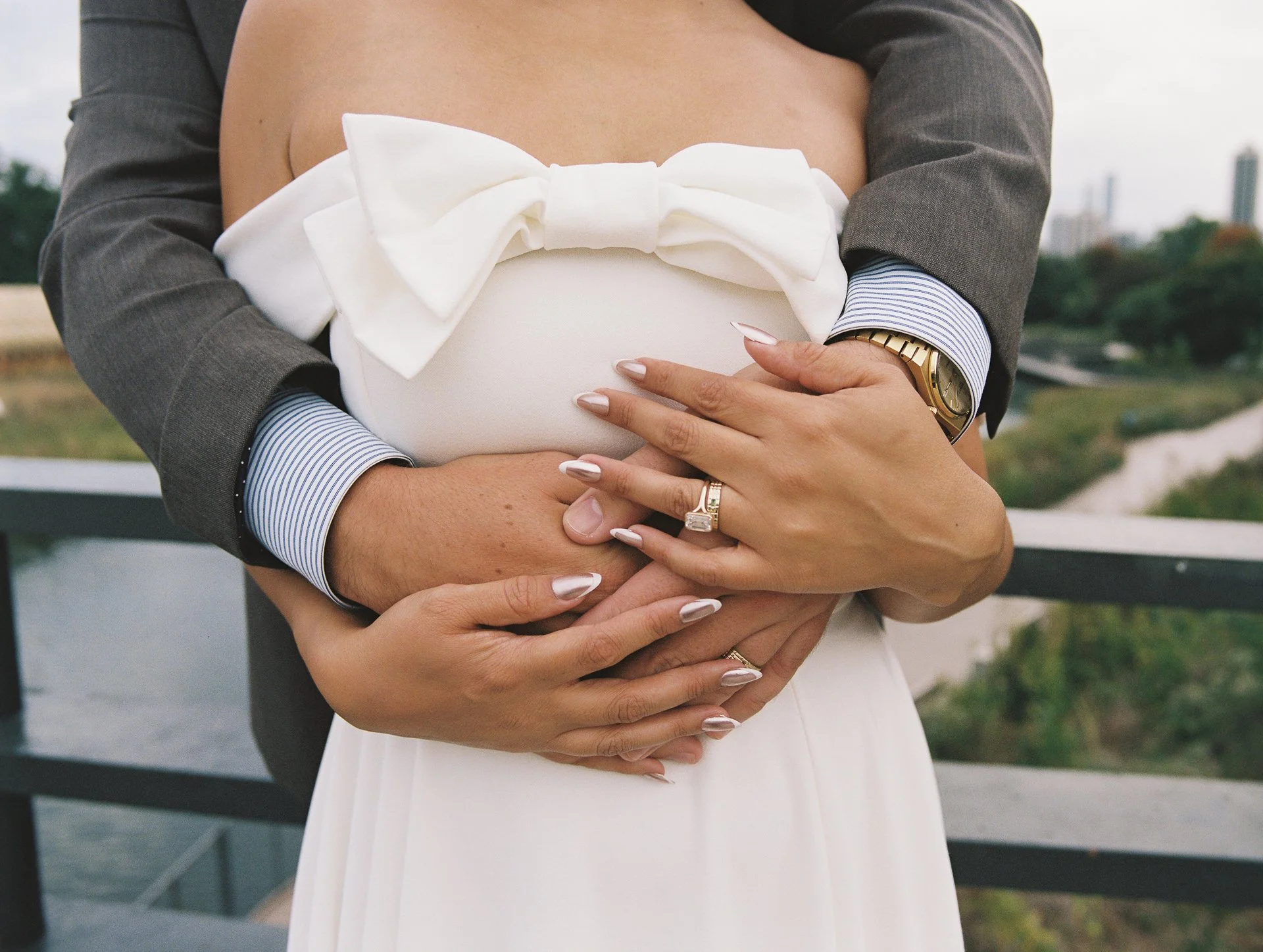 A close-up of a couple's hands and arms, with the woman wearing rings and the man wearing a watch, as they hold each other over the woman's pregnant belly, which is decorated with a large white bow.