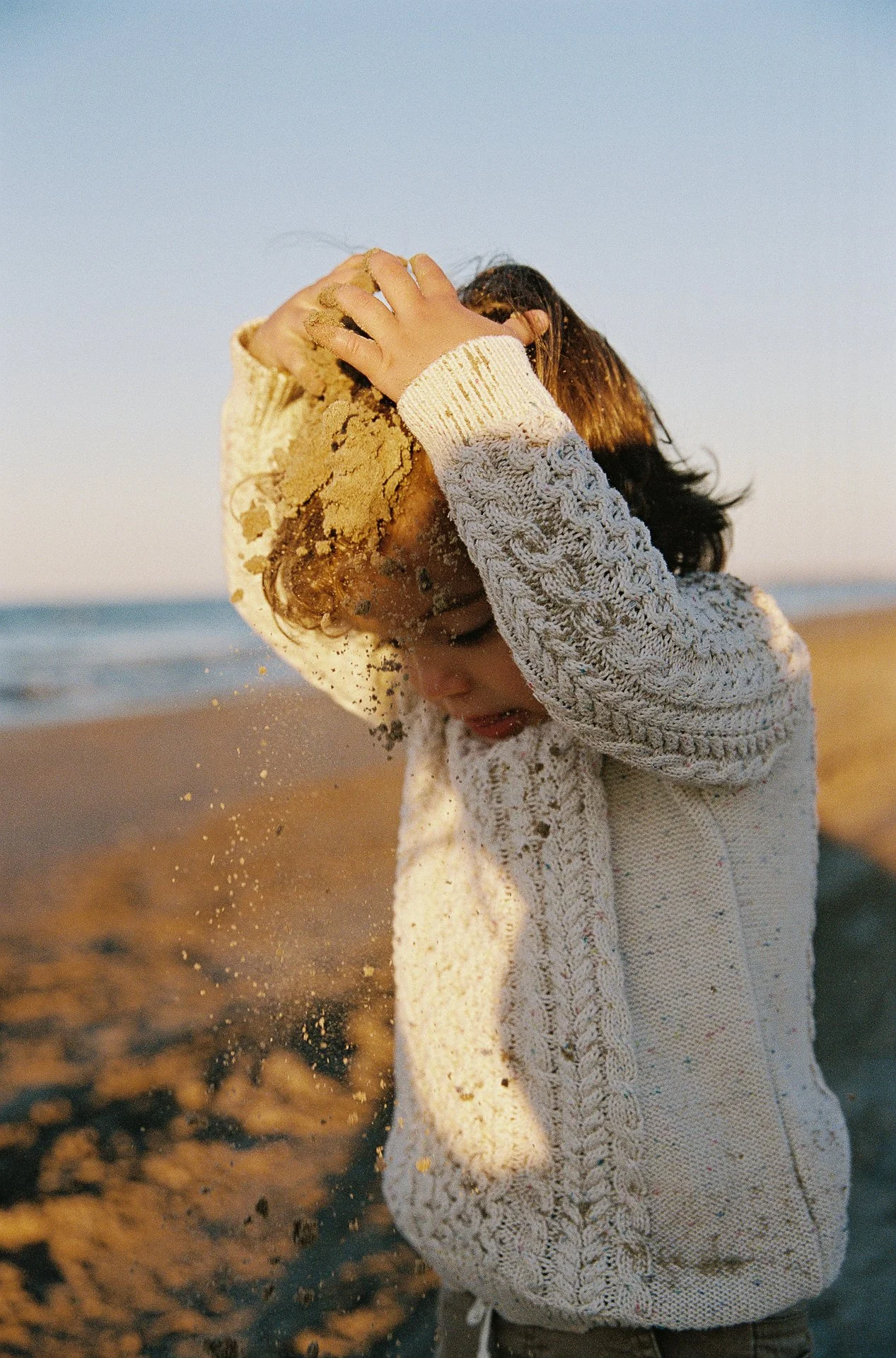 Child on beach wearing light-colored knit sweater, throwing sand on head, with ocean in background during sunset.