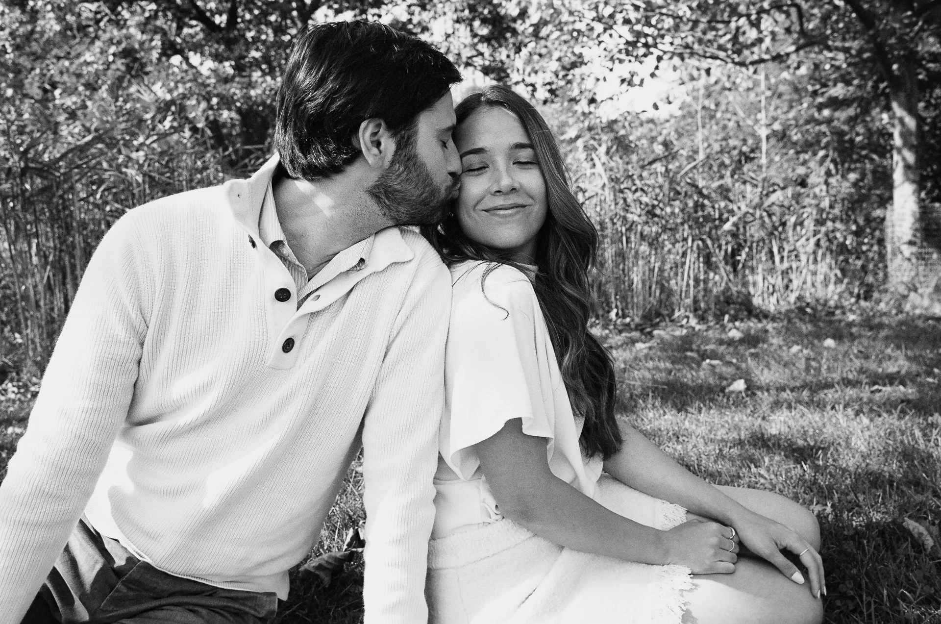 A black and white photo of a couple sitting outdoors on grass, with the man kissing the woman on the forehead and the woman smiling with her eyes closed.