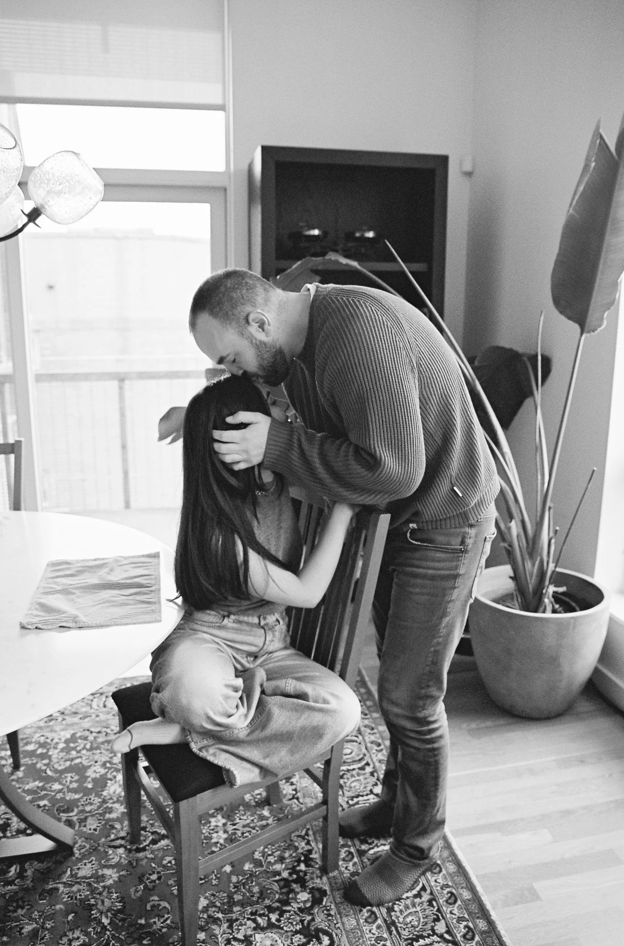 A man leaning over and kissing a young girl who is sitting on a chair with her eyes closed, in a domestic setting with a dining table, a potted plant, and a television.